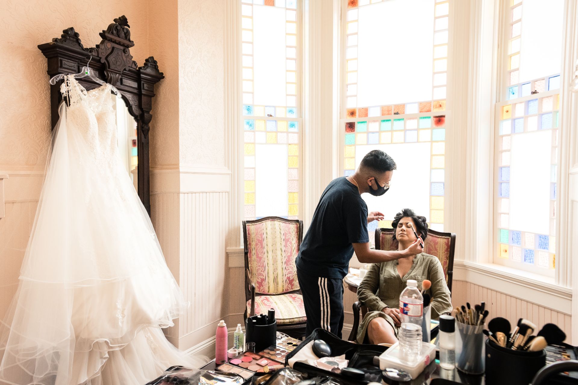 Bride is sitting in chair while makeup artist puts makeup on the bride. The brides dress is hanging in the background with stained-glass windows all behind.