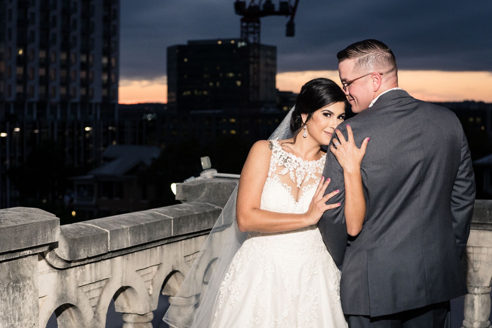 The bride is holding onto the arm of the groom. The couple is standing on a stone porch, the Austin downtown skyline is in the background.
