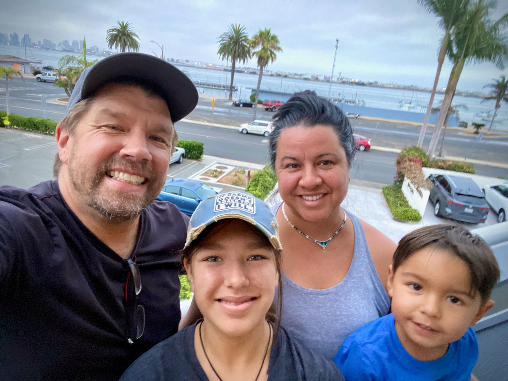 A family of four poses in a selfie photo. Father, Will Herrington, holds the camera and smiles with his wife and two children. There is a beach scenery off in the distant background.