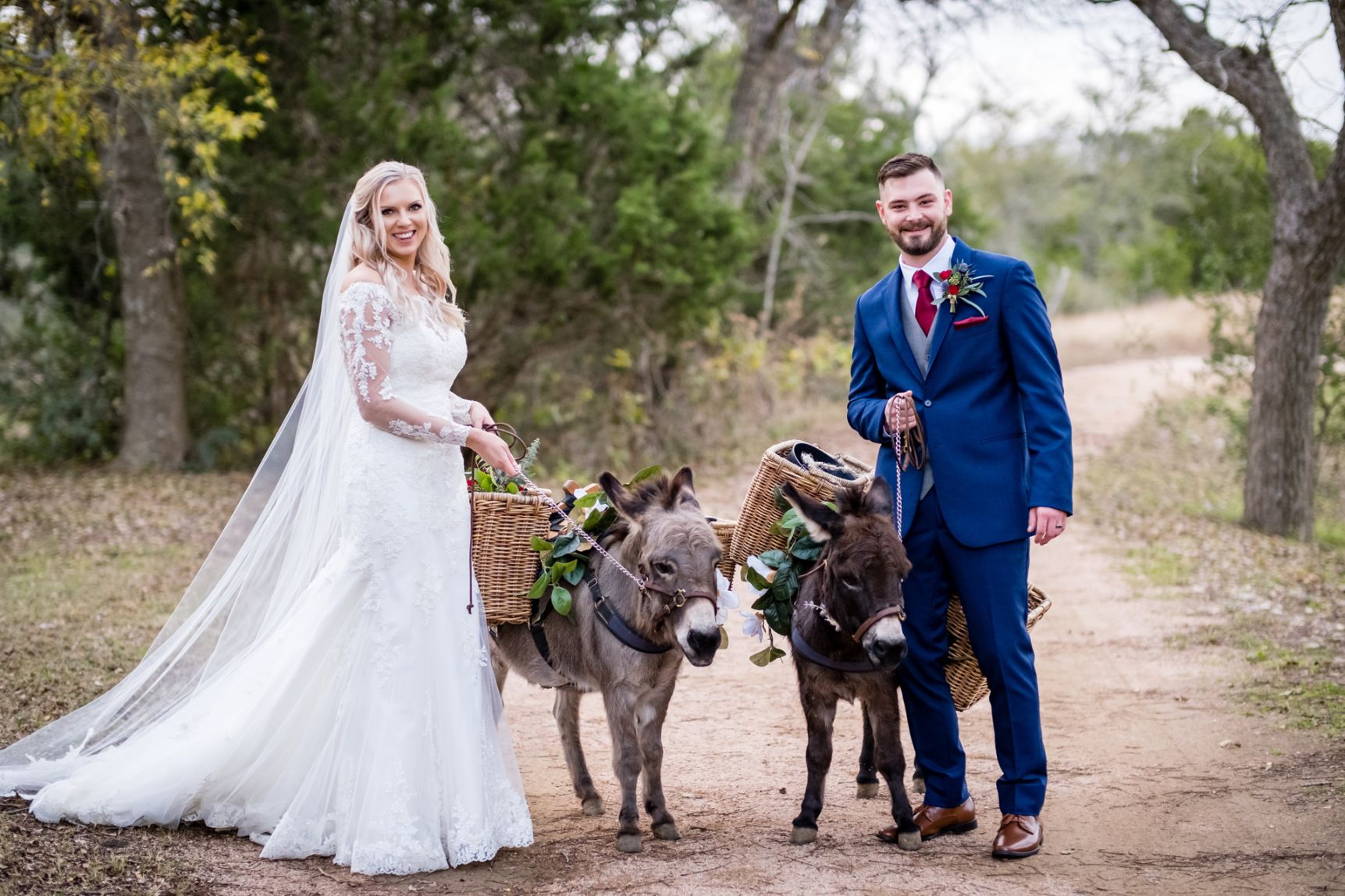Bide and groom stop to pose for a photo as they walk two donkies on a dirt path at Pecan Springs Ranch wedding venue. 