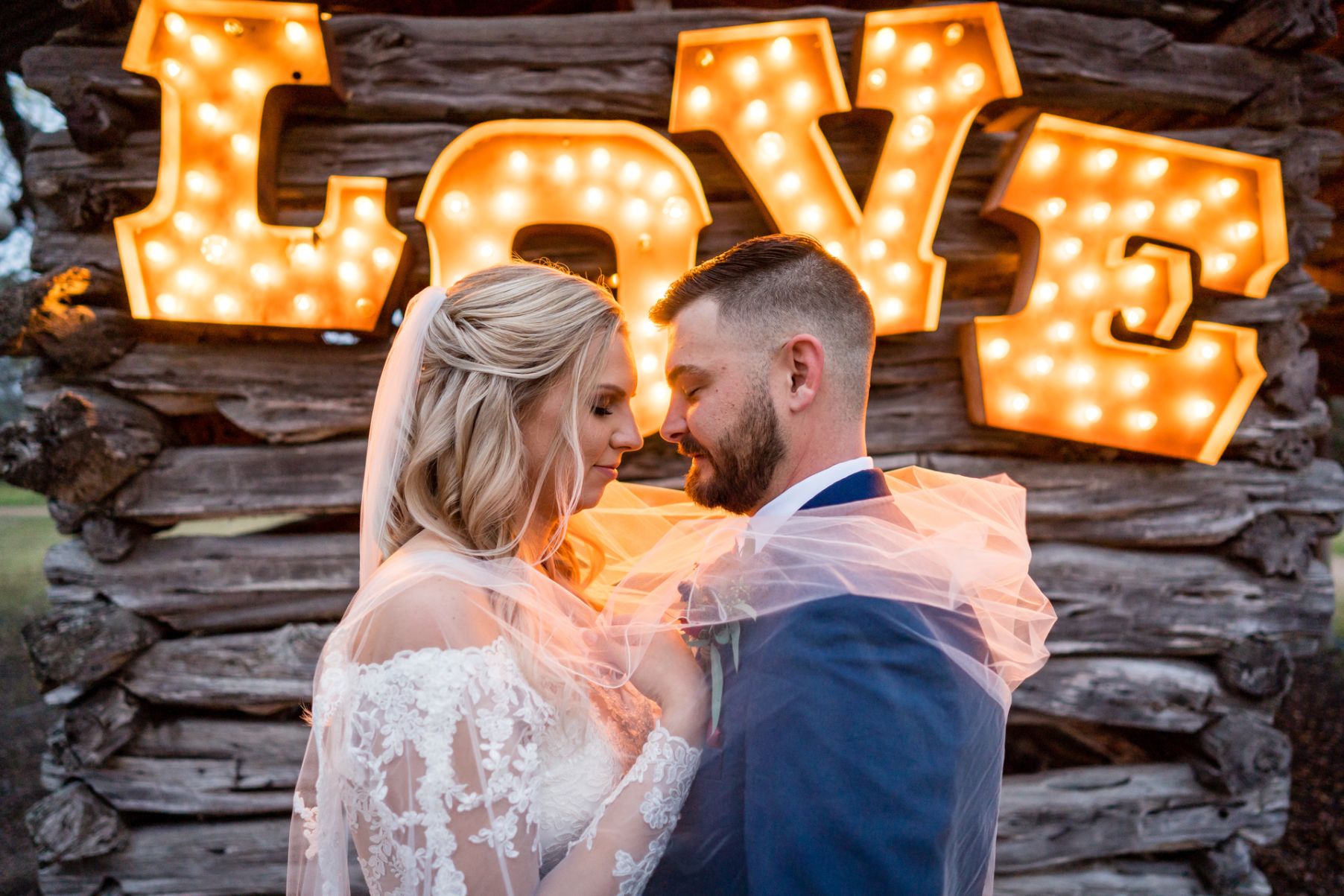 Bride and groom embrace closely holding each other with a light up sign that says love in the background of an old historic shack at Pecan Springs Ranch.