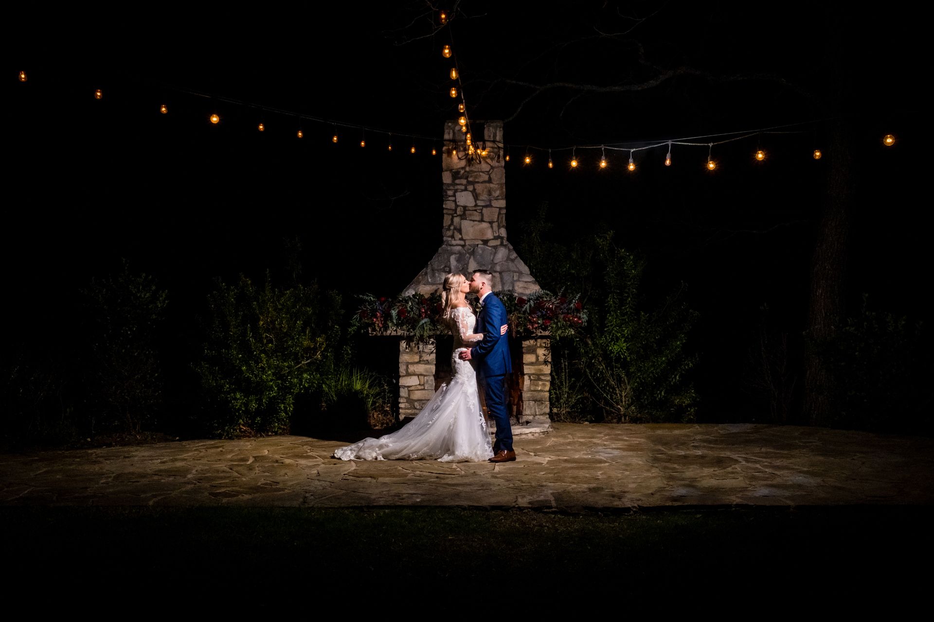 Bride and groom kiss under a string of bistro lights at night. A rustic fireplace is in the background. 