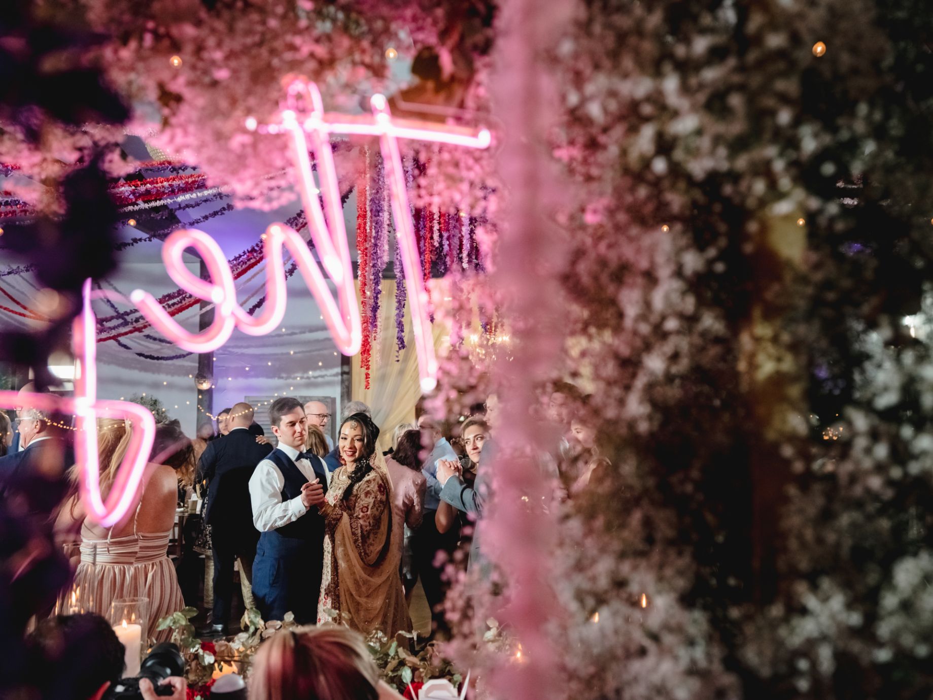 Brightly colored florals in pink, red and purple with a pink neon light is surrounding bride and groom dancing during their reception.