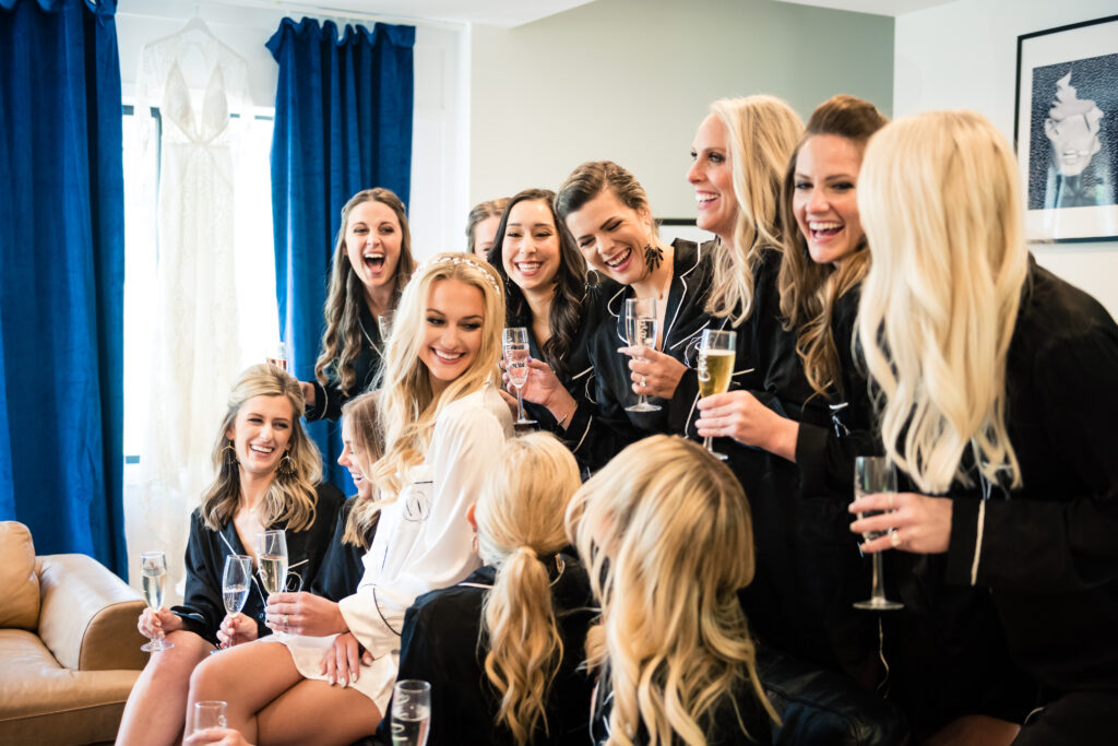 Bride is sitting on bed with 11 bridesmaids. Everyone is wearing matching, black silk pajamas and is smiling and laughing at each other, holding a glass of champagne.