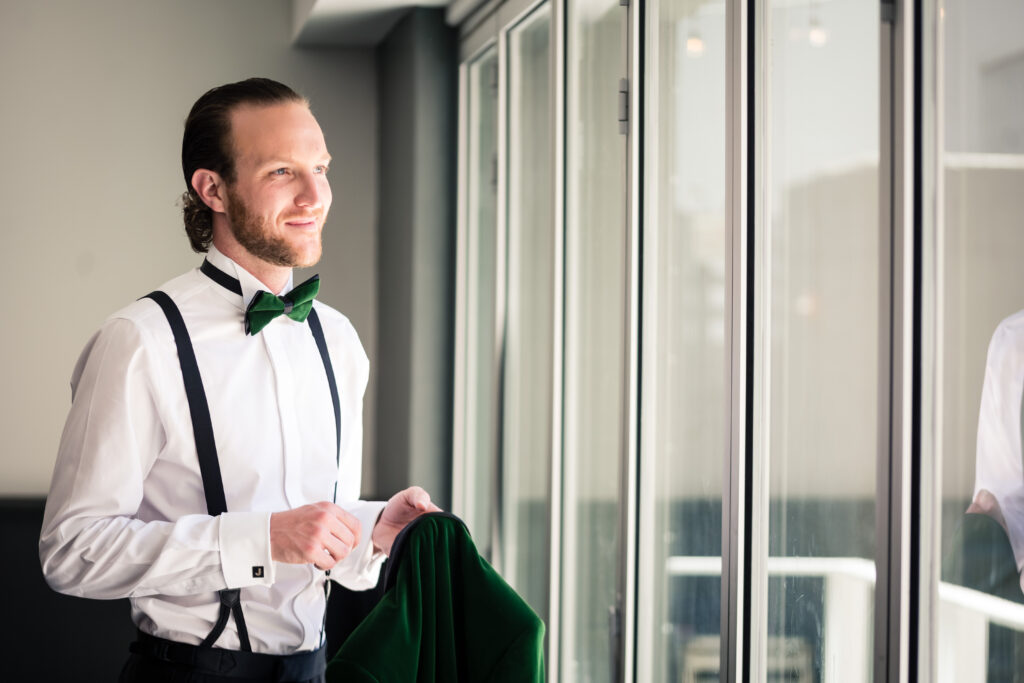 The groom in his white suit, black suspenders, and velvet green bow tie is holding a matching velvet green suit, looking out the glass windows, smiling.