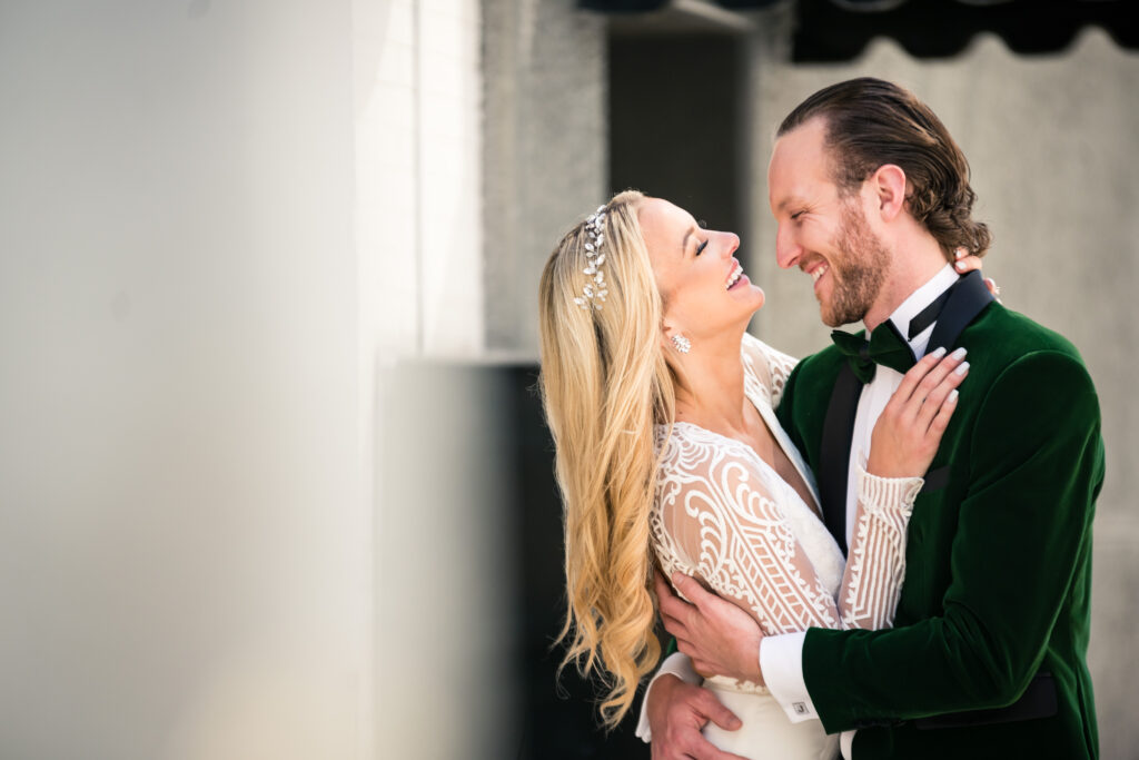 Bride and groom face each other, laughing. Bride has long blonde curly hair and is wearing a white lace gown with bead headband. The groom is wearing a unique green velvet suit. 