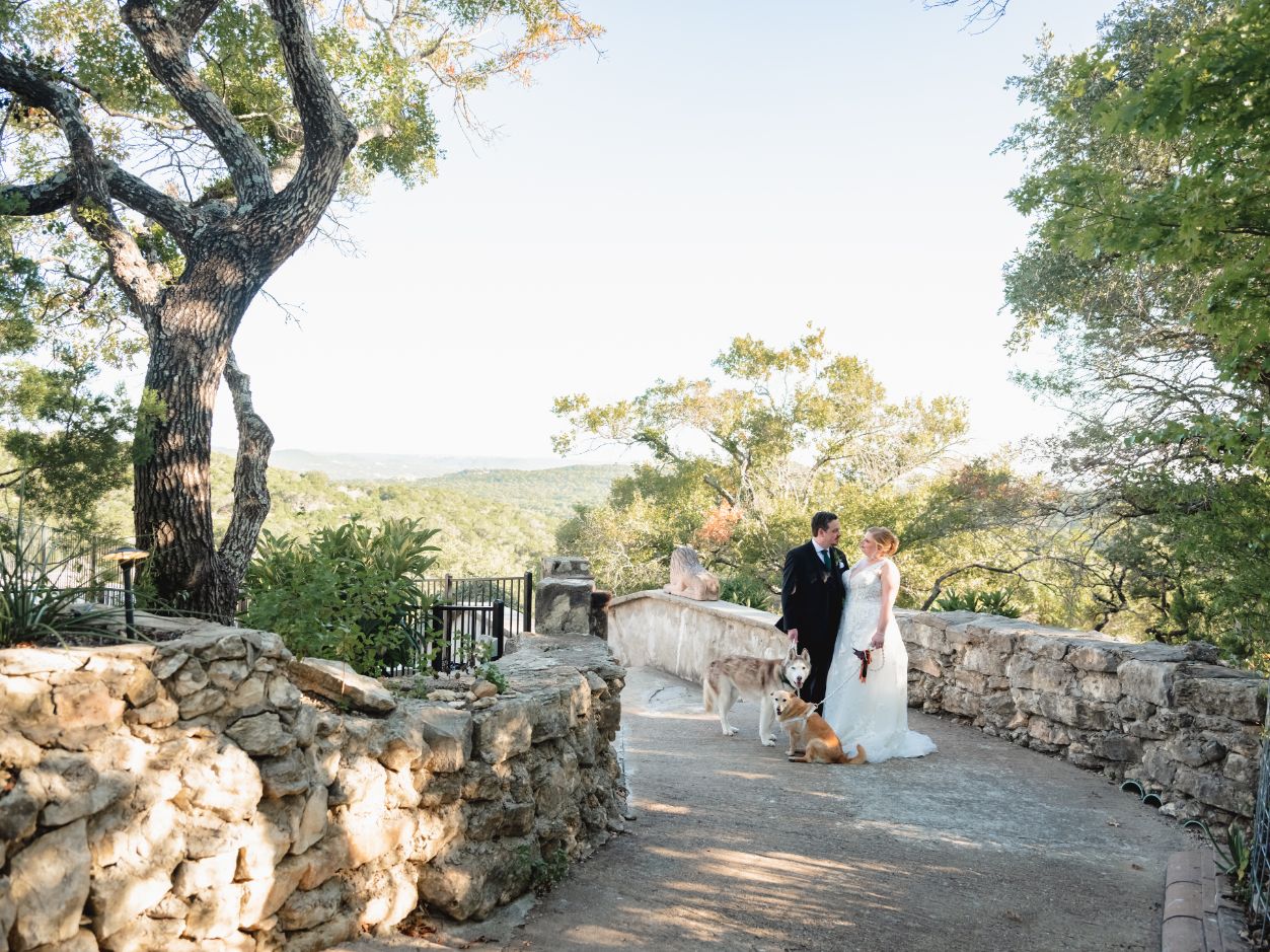 Bride and groom look at each other, holding leashes for two dogs. A rock-lined pathway leads out to a scenic view of green rolling hills.