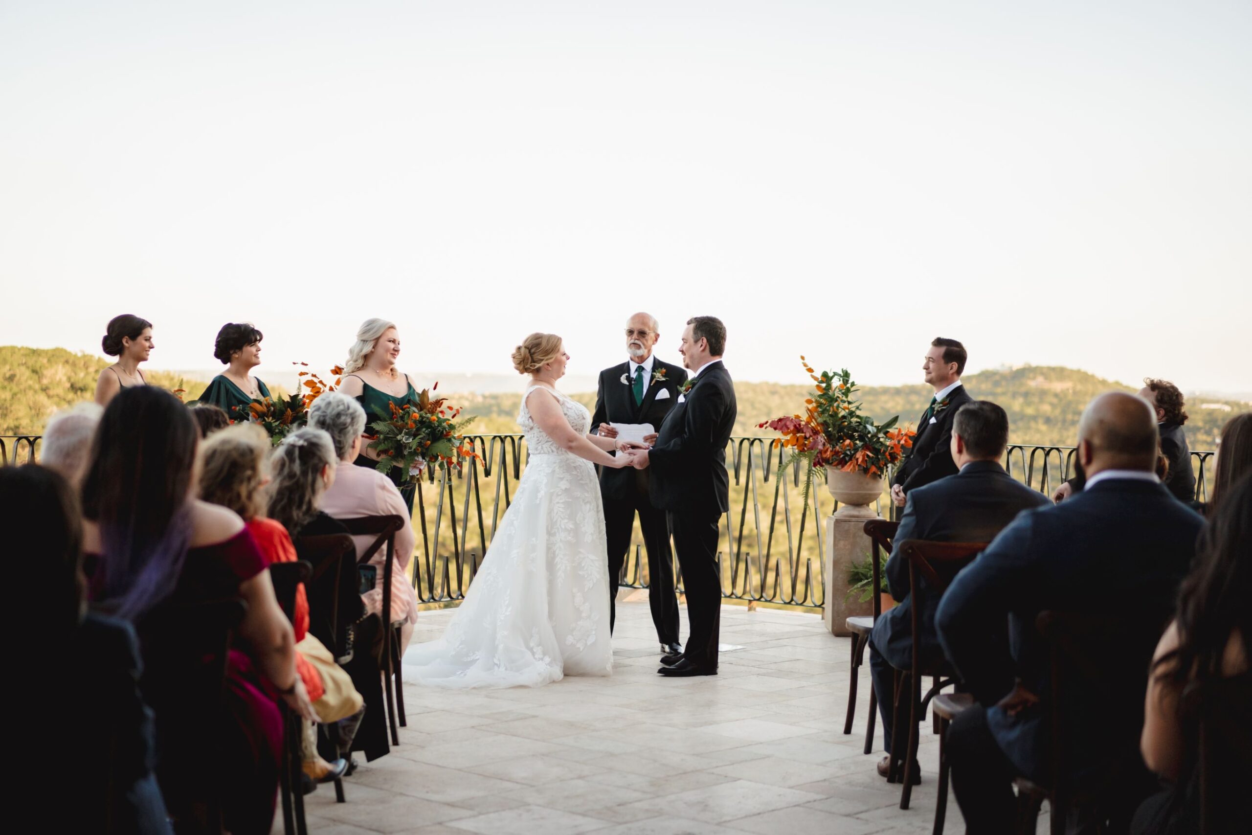Bride and groom hold hands, looking at each other during wedding ceremony. Officiant stand behind them with olive green hills in the background. 