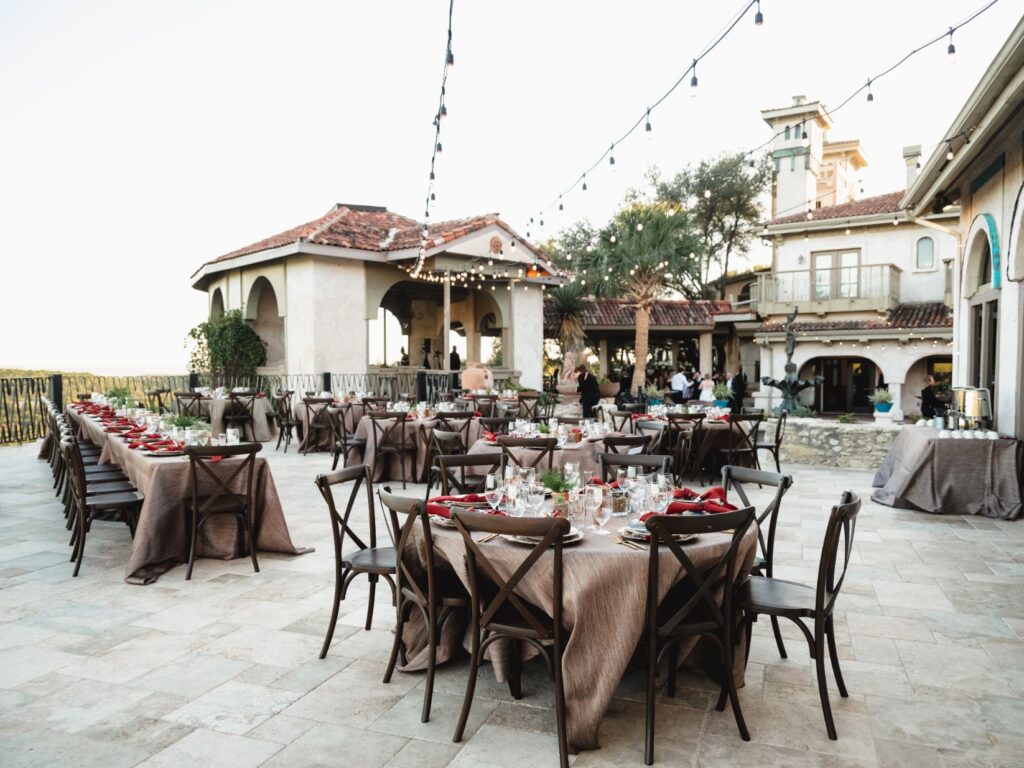 Tables are set with brown wood chair and red napkins on a patio outdoors with bistro lights hanging above and the building in the distance at Villa Antonia wedding venue. 