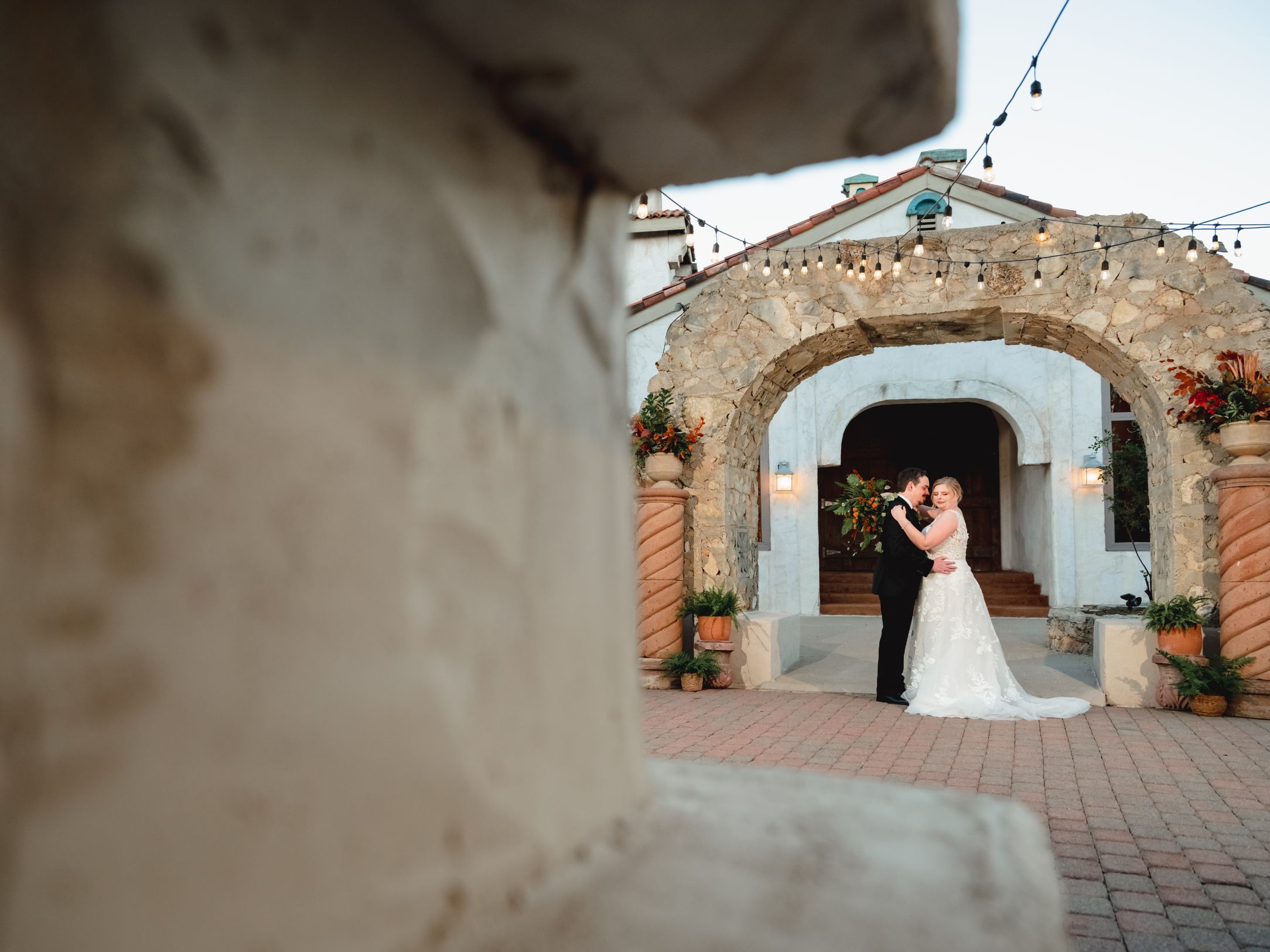 Bride and groom hold each other under a stone arch with stone pillars on each side and bistro light hanging above.