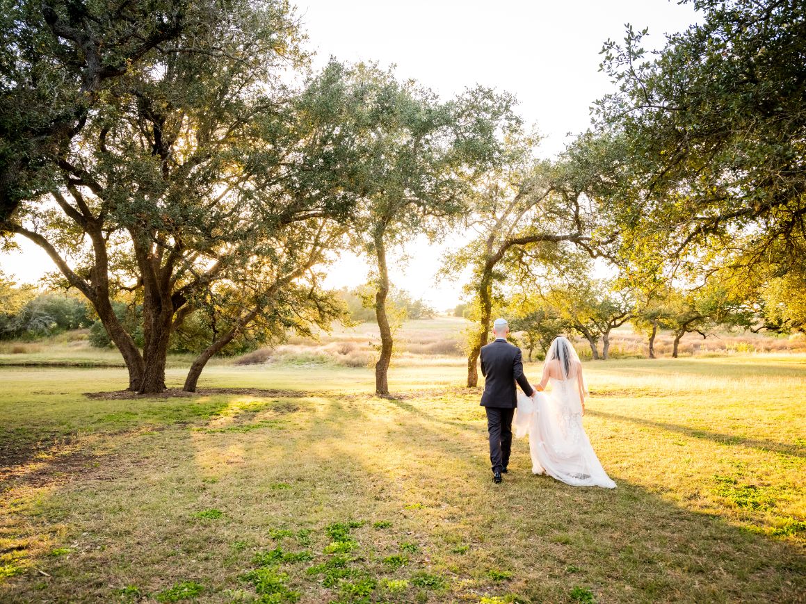 Groom and bride walk into the distance with sunset rays poking through the oak trees.