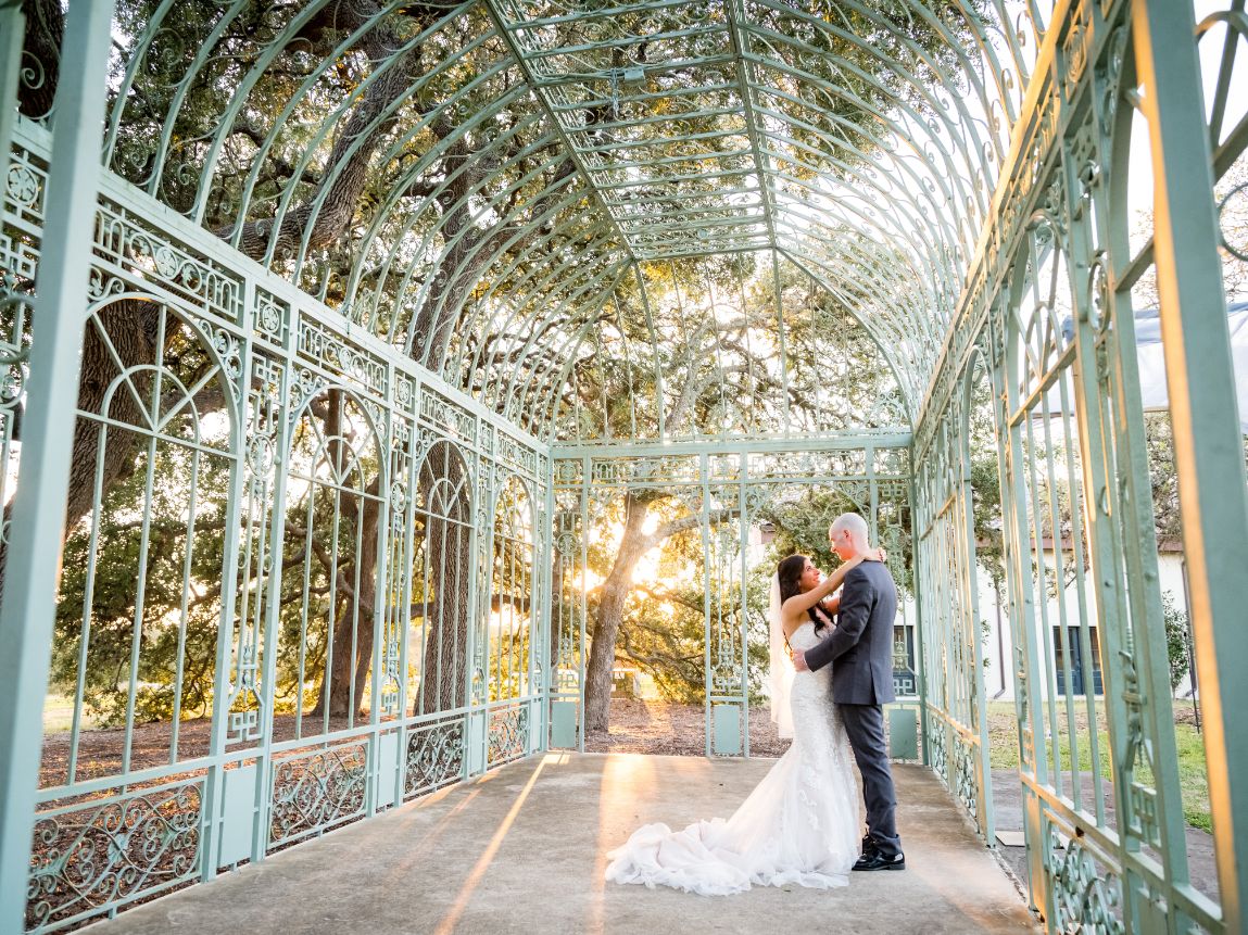 Bride and groom hug underneath a French-style green gazebo with warm sunlight poking through the Oak trees in the background.