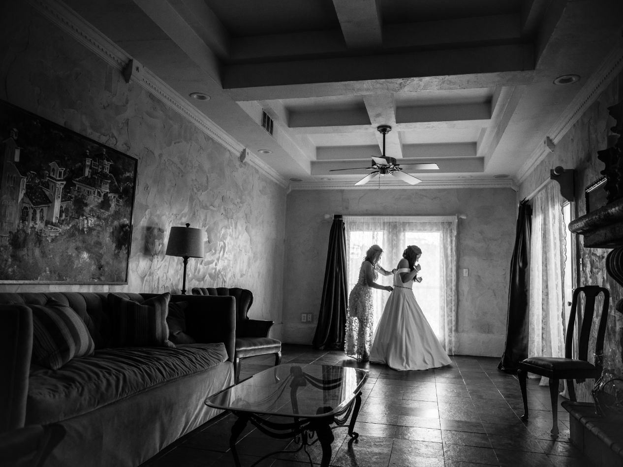 The bride is standing in a European style room wearing a white wedding dress. The bride's mother is helping zip up her dress near a window at Villa Antonia wedding venue.