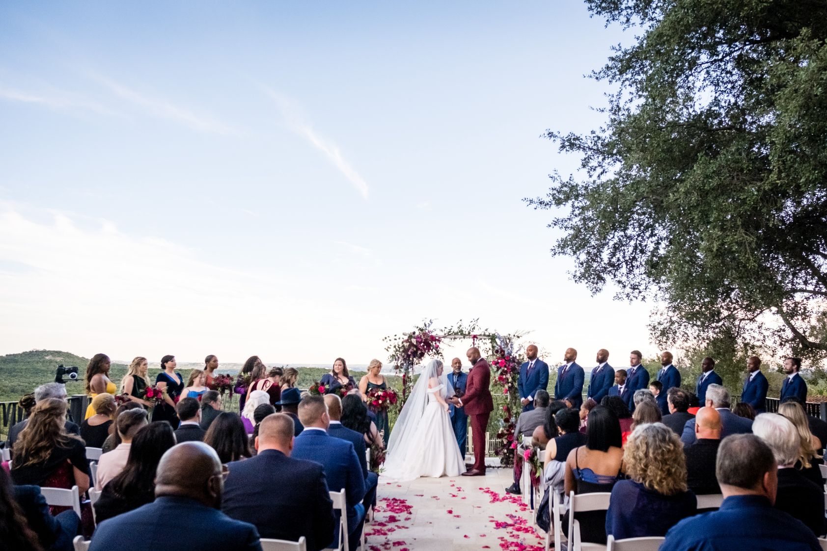 Bride and room hold hands at floral arch altar. Red floral petals are scattered on the ground. The bridal party is lined up on each side of the couple, and the wedding guests are sitting in rows of white chairs. Clear blue skies and green rolling hills are in the background.