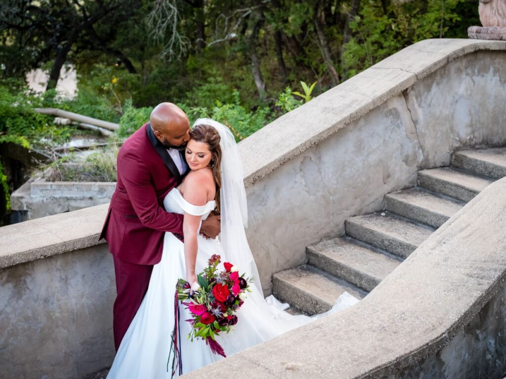 The groom wearing a maroon suit is holding the bride, kissing her forehead as they both stand on a stone stairway. Green trees and foliage is seen in the background of Villa Antonia wedding venue. 