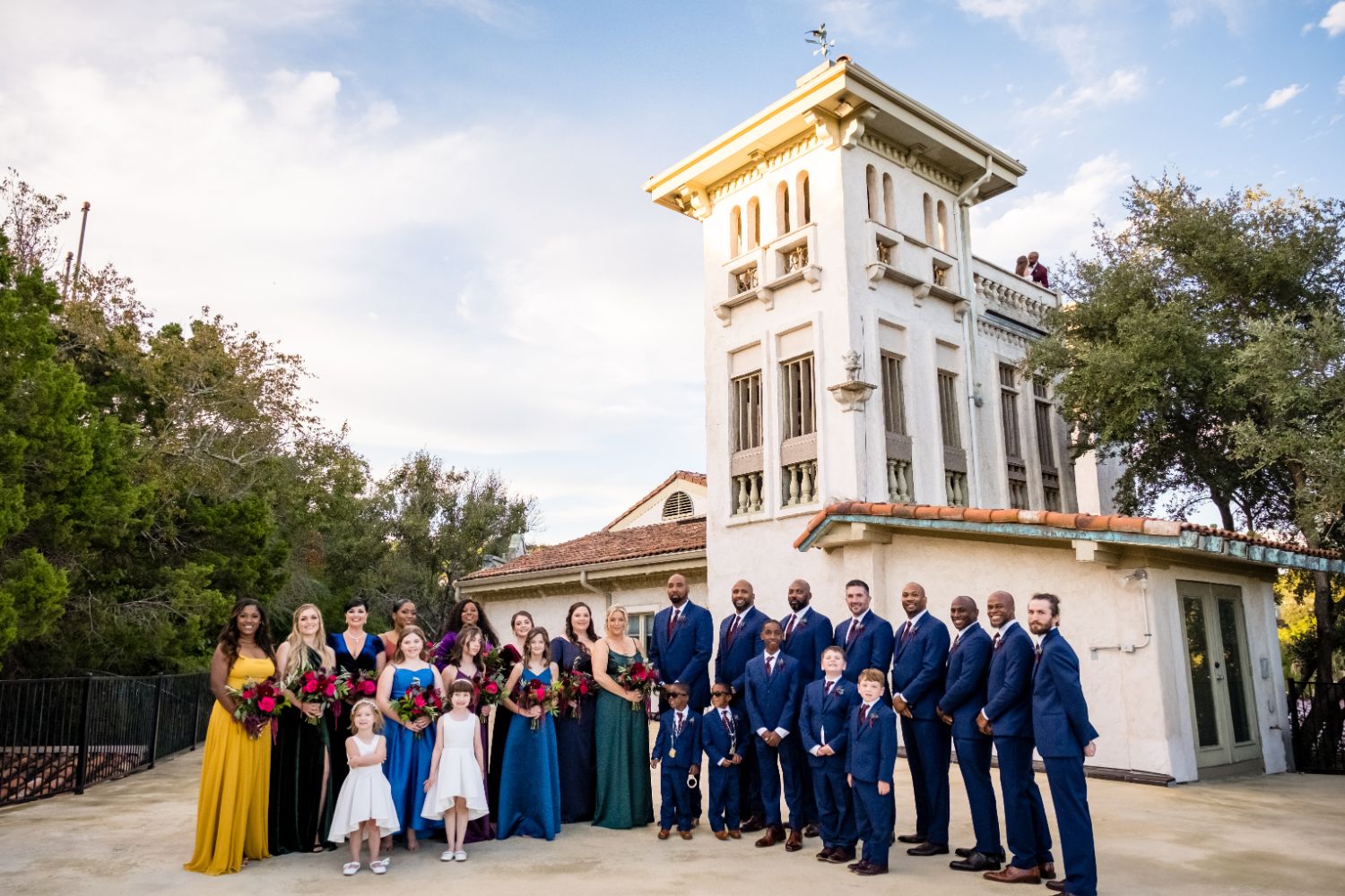  A bridal party of 26 people are standing side by side in front of a large tower building with Tuscany architecture at Villa Antonia wedding venue. 