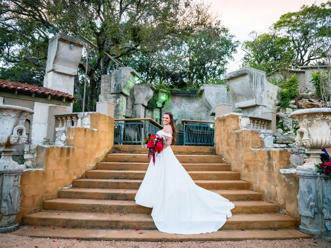 The bride is standing on a stone stairway in a white dress, holding a red bouquet of flowers.  Trees and large stone pots and fountain is in the background. 