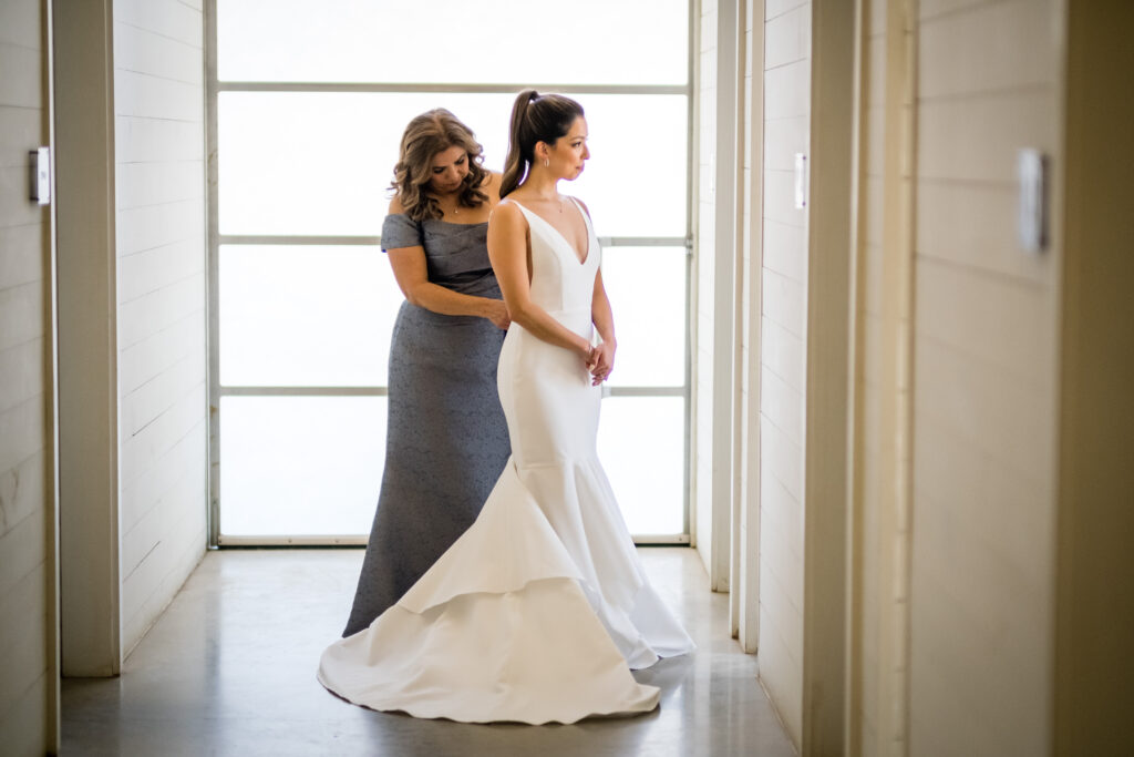 The bride's wedding gown is zipped by her mother in the hallway of the Prospect House, the bright light from outside goes through the window behind them.