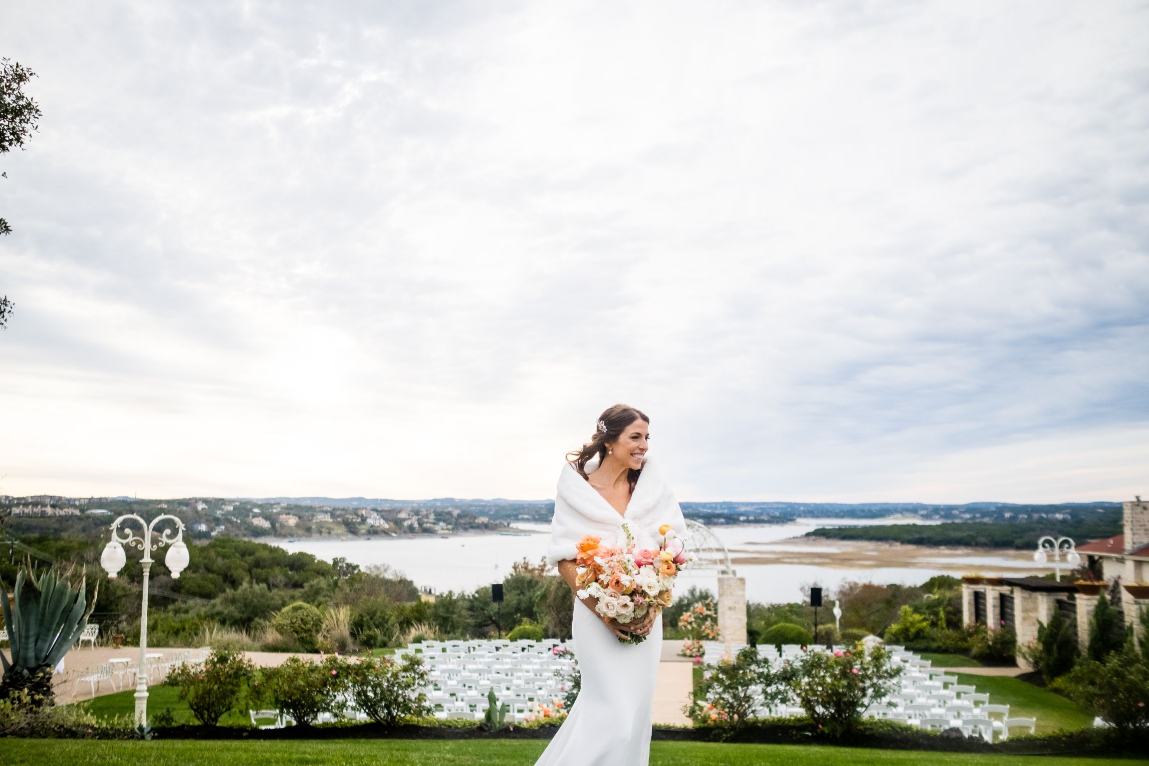 A bride wearing a white dress and fur cover smiles while holding a bouquet of pink and orange flowers. Lake Travis is seen in the distance.