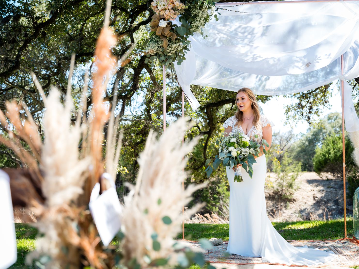 Bridre is holding a bouquet of white and green flowers as she stands under and wedding altar with a white sheet blowing in the wind above her. 