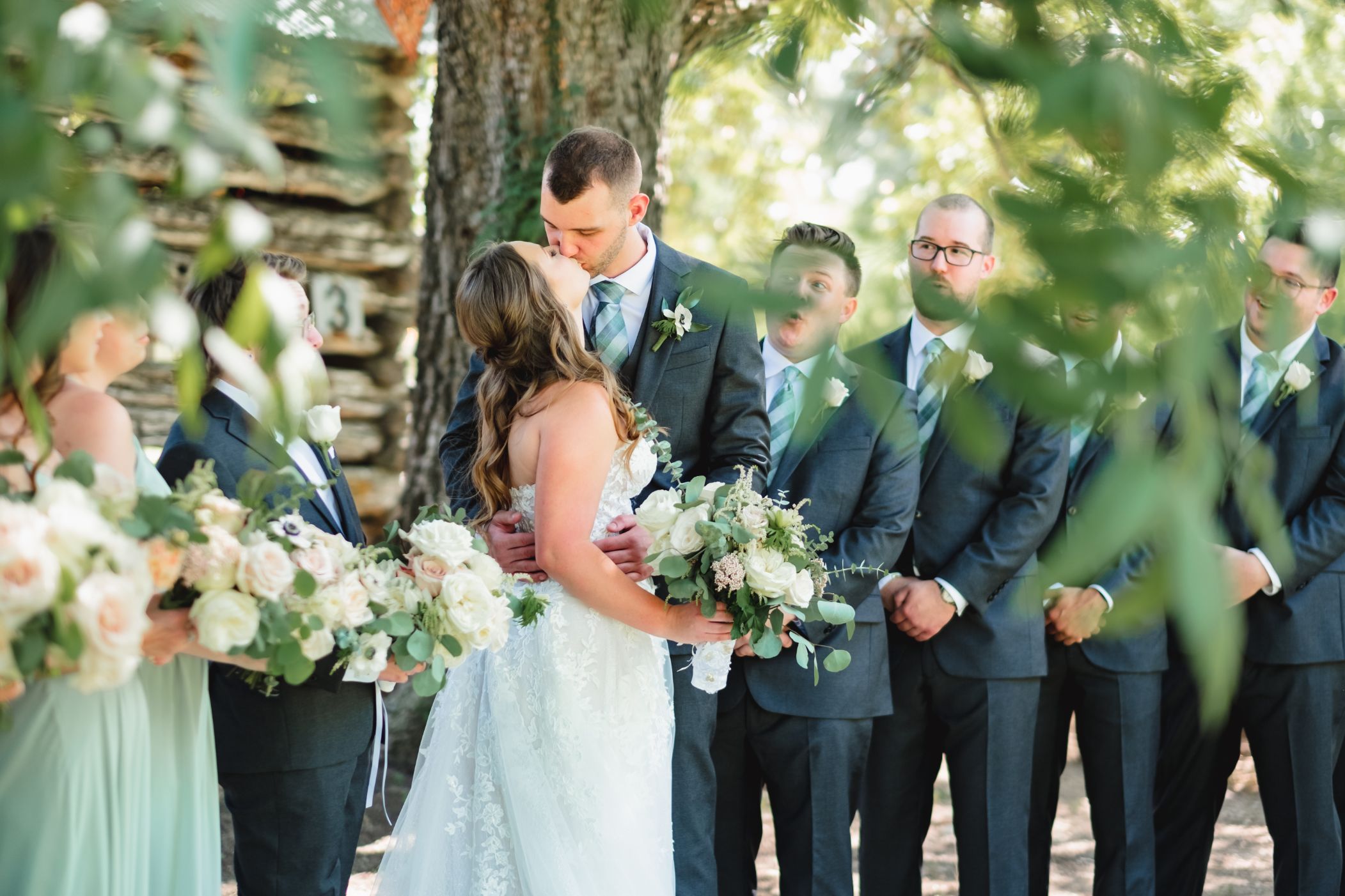 Groom and bride kiss under a large green tree. Bridal party is looking in excitement for the couple.