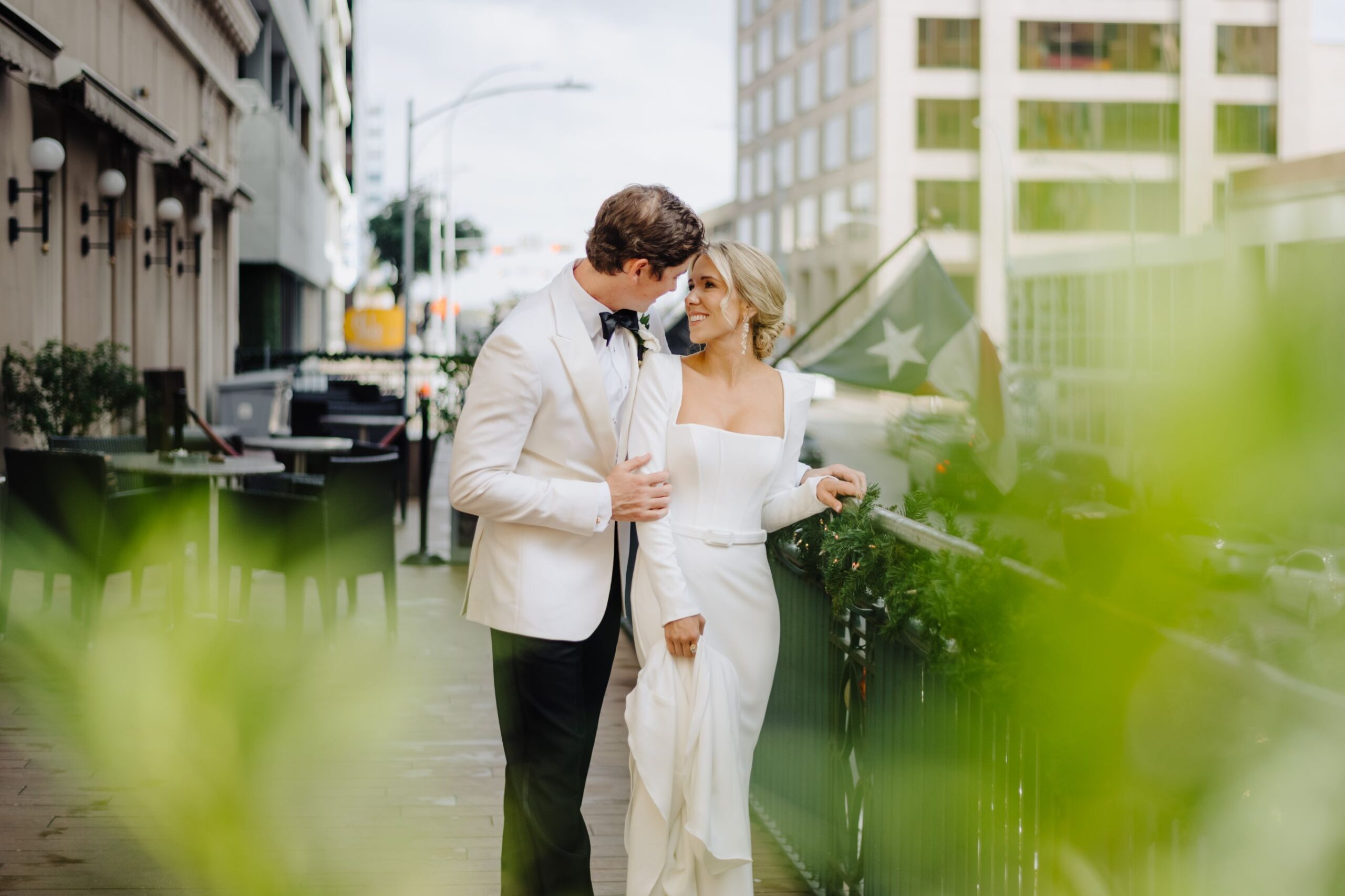 Bride and groom are walking on a patio, smiling and looking into each other's eyes. A Texas flag hangs in the background with tall Austin buildings in the distance.