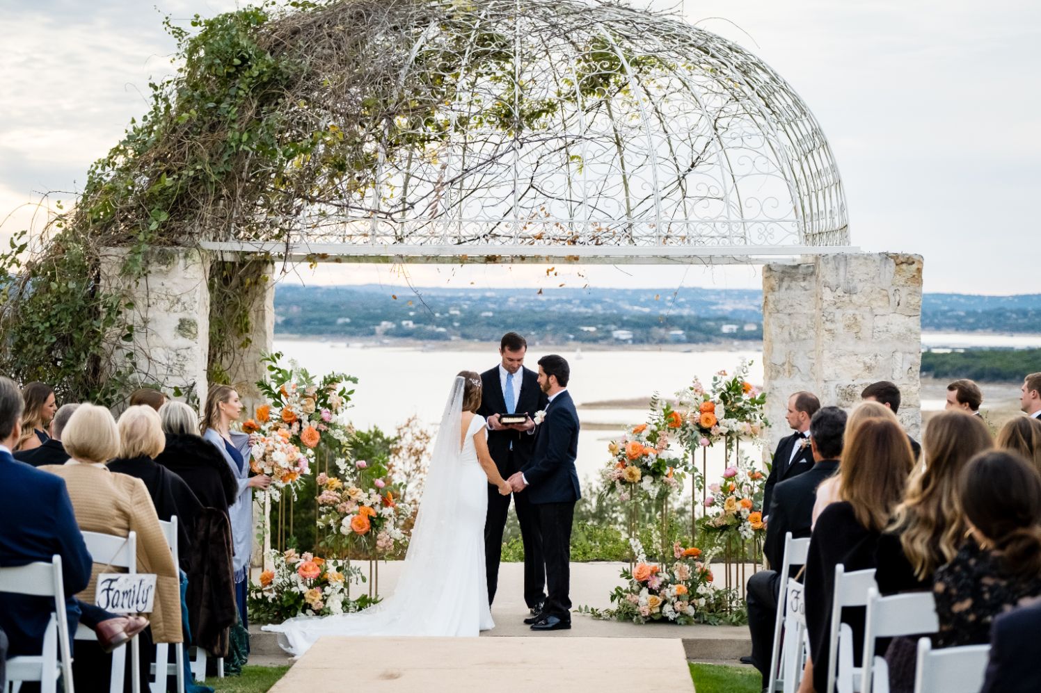 Bride and groom hold hands at the altar of their wedding ceremony at Vintage Villas in Austin, Texas. Wedding guest sit in white chairs and an officiant is speaking behind the couple. Lake Travis can be seen in the distance.