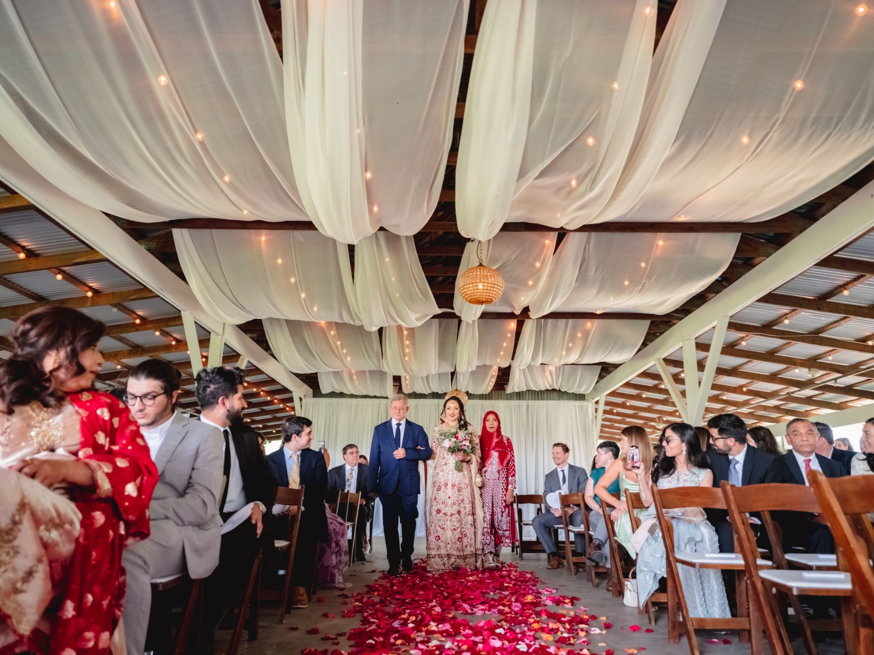A bride wearing ethnic wedding garments is walking down the ceremony aisle with red flowers on the floor at Pecan Spring Ranch wedding venue. Guest are seated in brown wood chairs. 