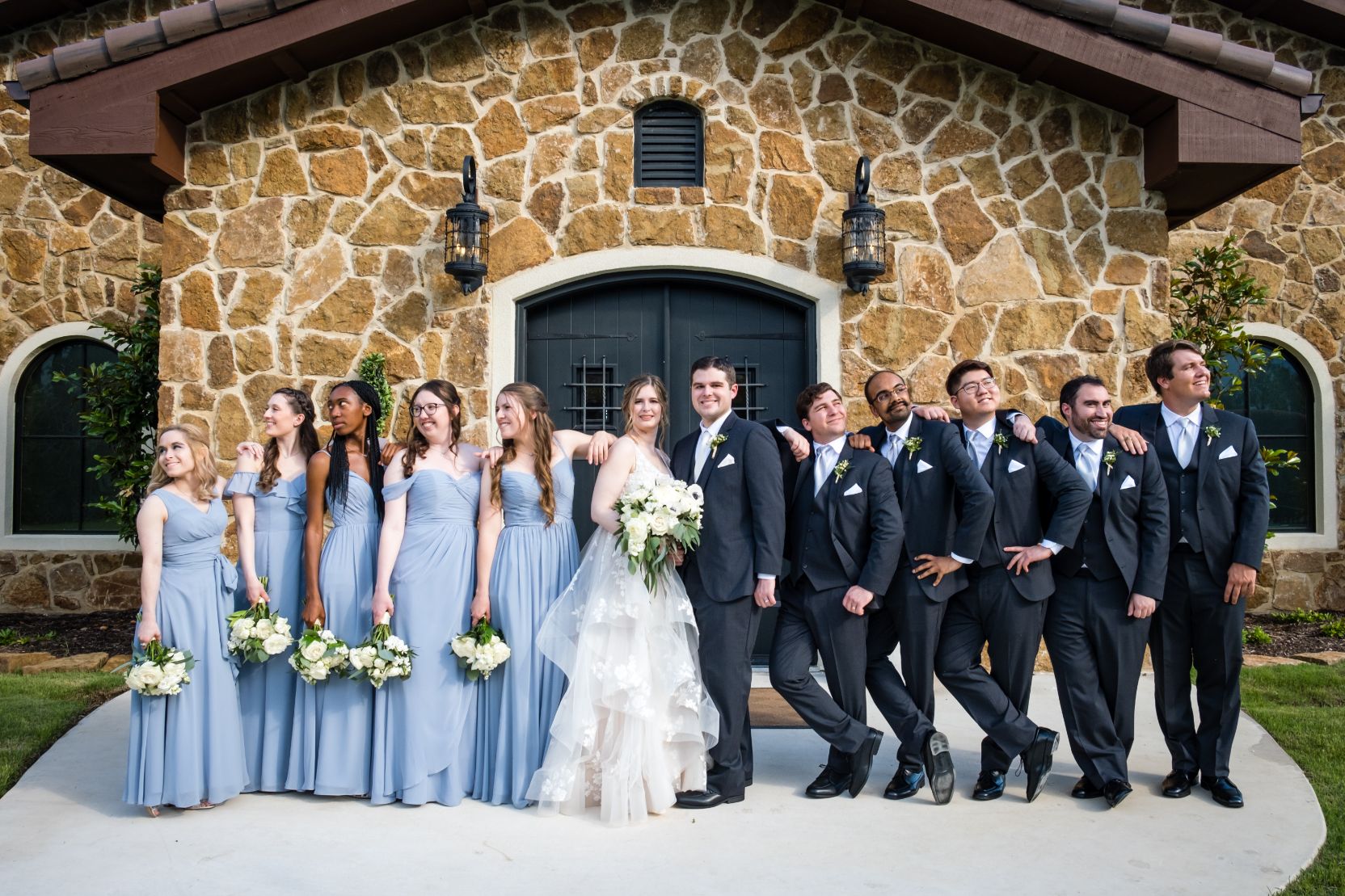 A bridal party of 12 people stand in front of a rock faced building with large lanterns and black double doors. 