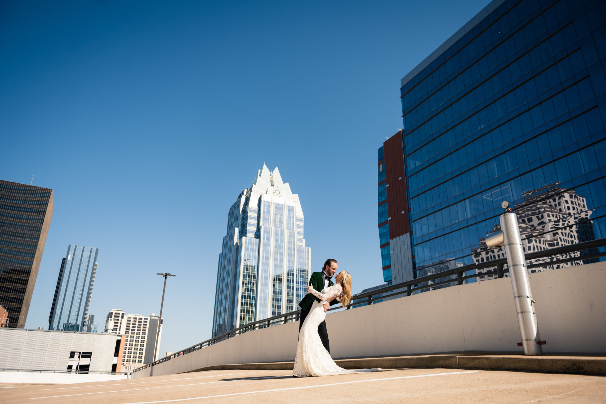 A groom wearing a green velvet suit dips his bride while kissing her. Austin, Texas skyline can be seen in the distance.