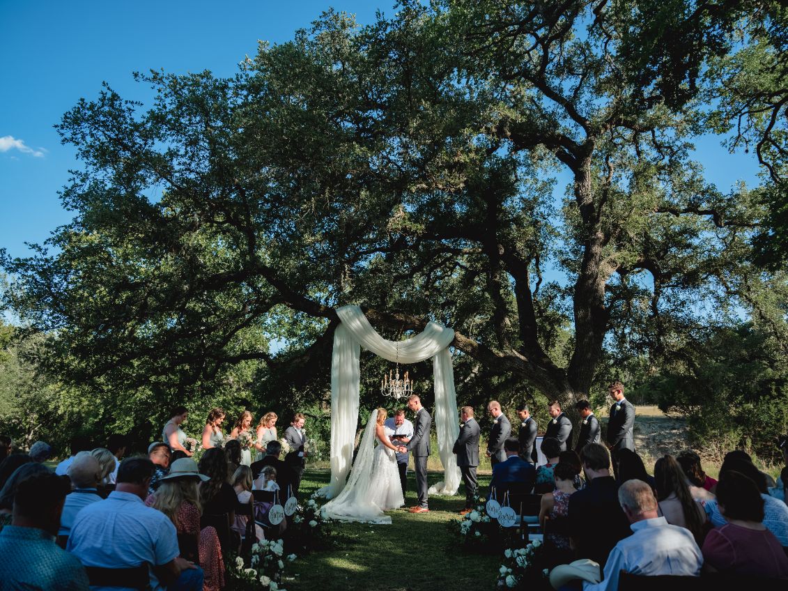 Bride and groom hold hands under a white fabric arch hanging from large tress above during the wedding ceremony at Pecan Springs Ranch. 