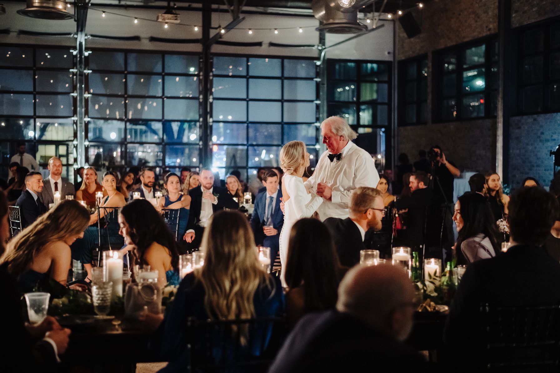 Bride dances with her father during a wedding reception at 800 Congress. The guests are seated at tables and a large wall of glass windows are in the background. 