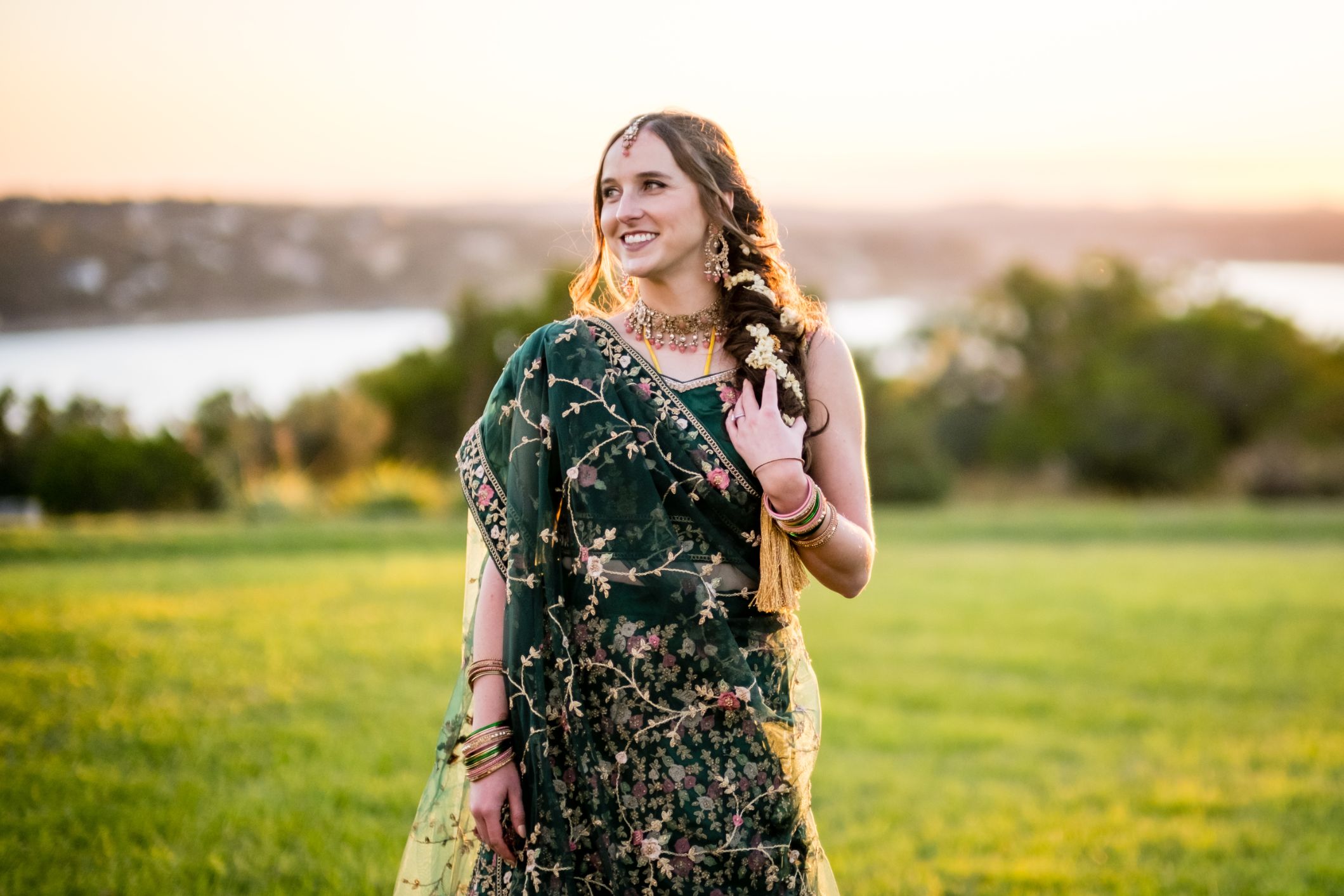 A bride wearing traditional Indian garments plays with hair and looks into the distance. Green trees, Lake Travis and a warm sunset are in the distance. 