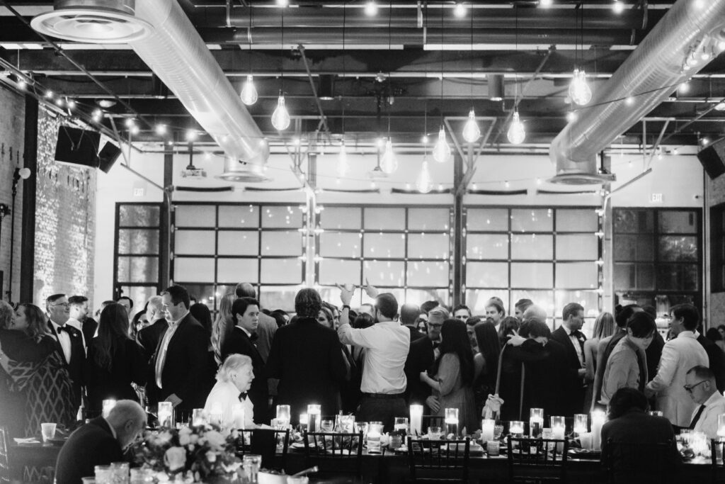 People attending a wedding at 800 Congress are standing and conversing. Tables at the venue are set with romantic candlelights in water vases. A large wall with windows and exposed, industrial beams can be seen around the venue. 