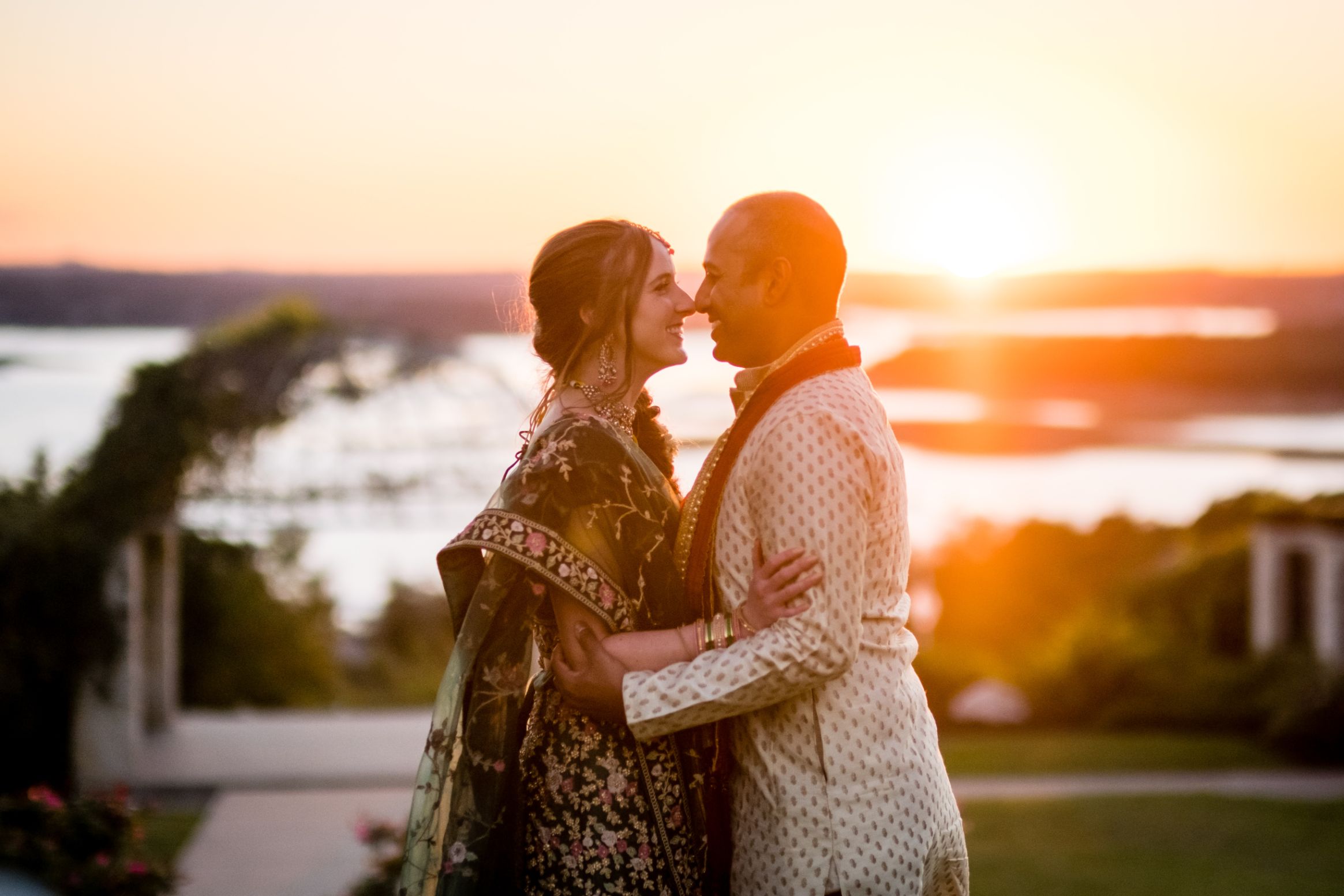 Bride and groom wearing traditional Indian garments look into each other's eyes with noses touching. An orange and yellow sunset with views of Lake Travis are in the background of Vintage Villas wedding venue.