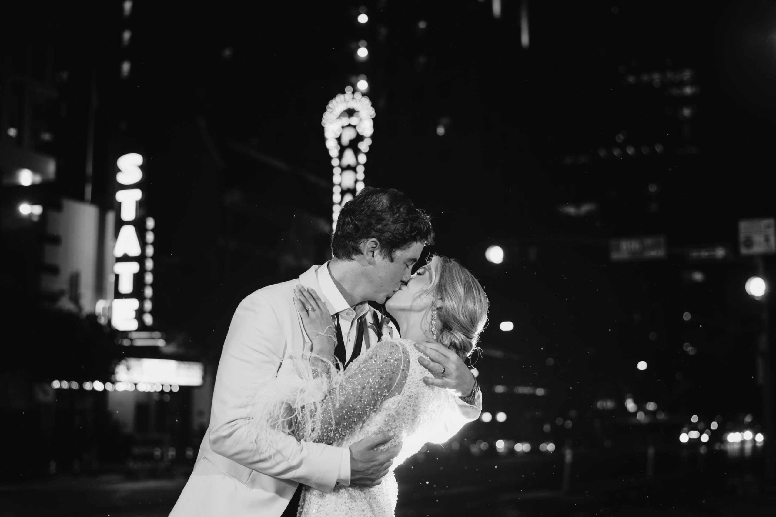 Groom and bride kiss at night, with the city lights and signs of Austin, Texas blurry in the background. 
