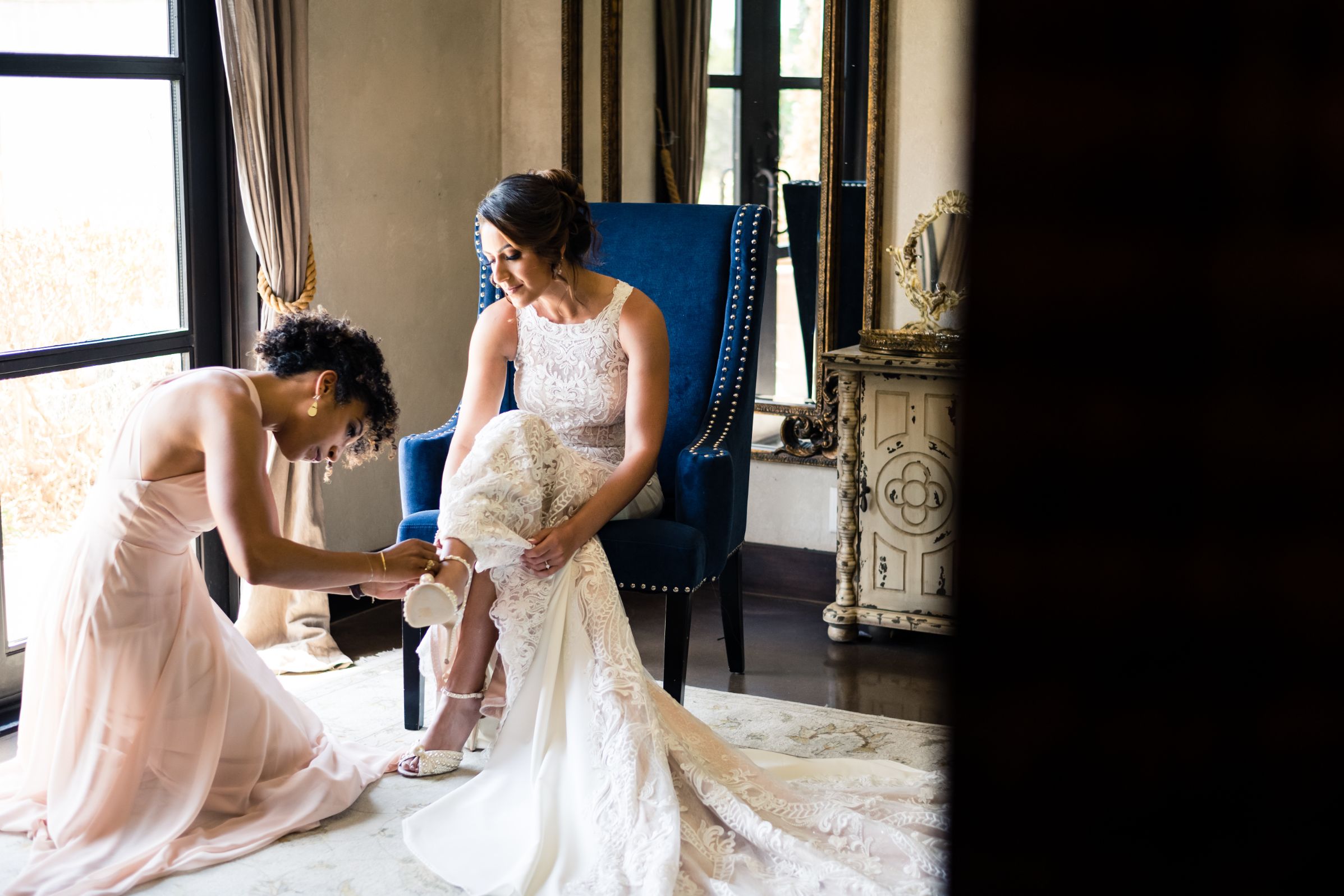 A bride sits in a blue chair while a bridesmaid kneels to help the bride buckle her shoes in a bridal suite.