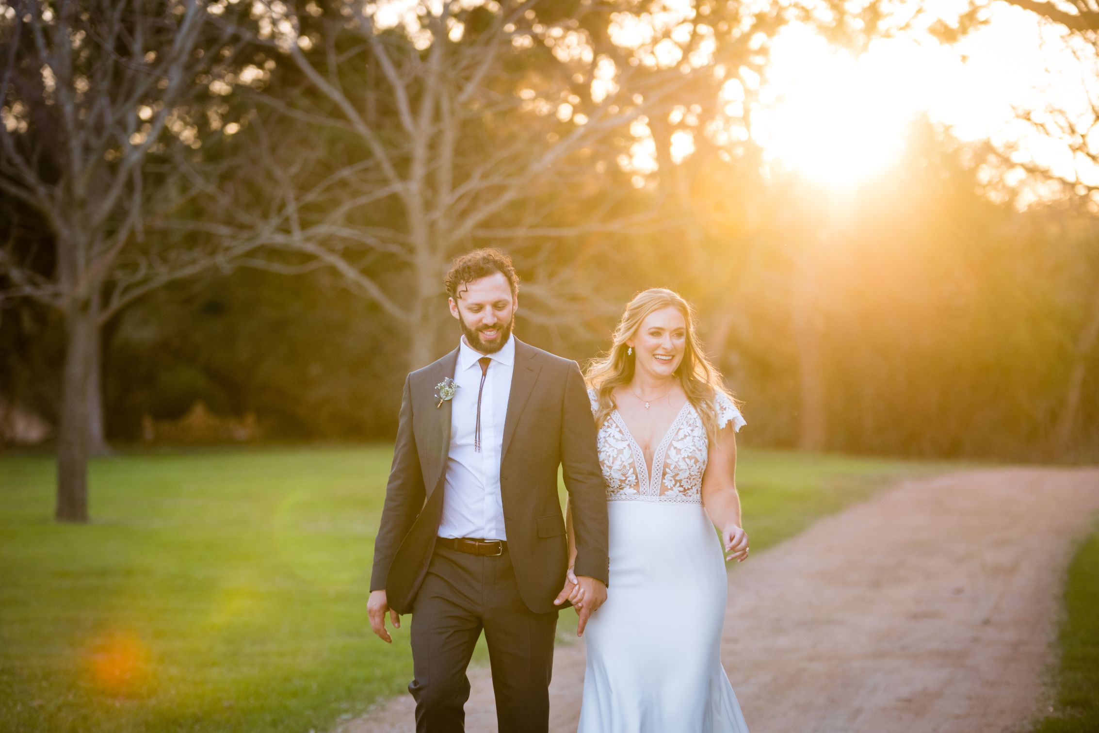 Bride and groom hold hands walking down dirt path with trees and a large warm sunset in the background.