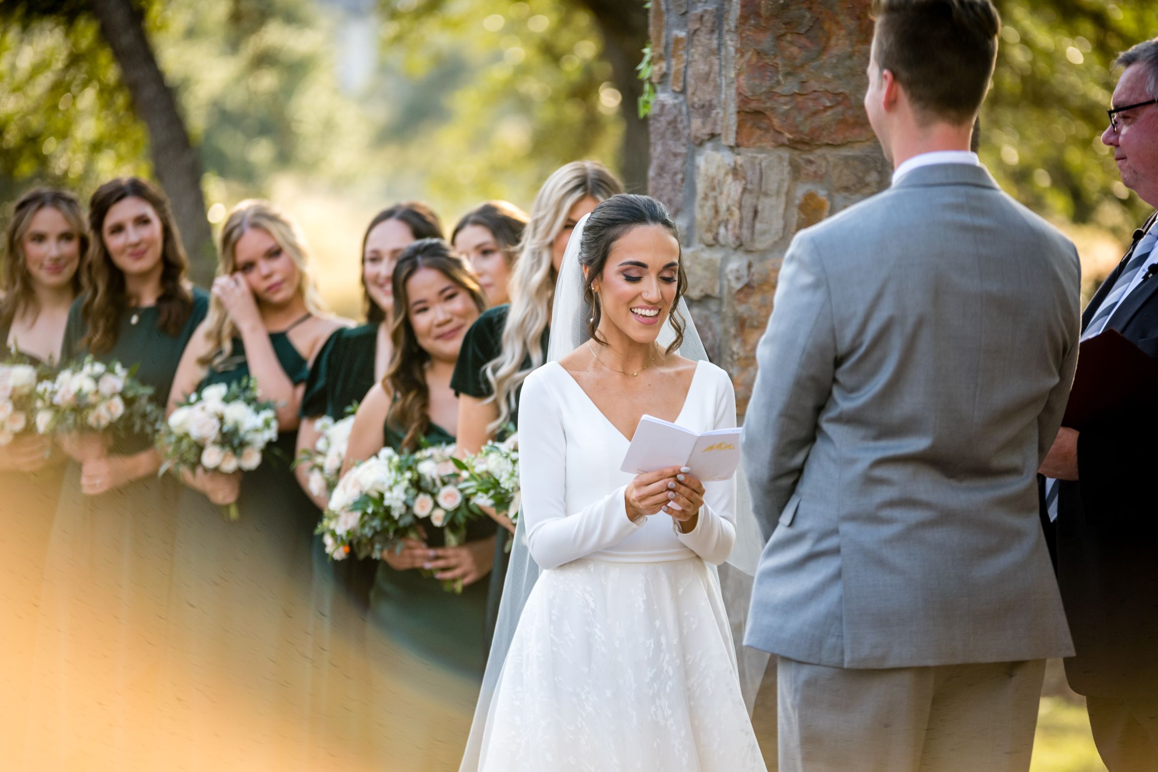 Bride reads off vow card to her groom standing in front of her with bridesmaids in green dresses lined up in the distancing smiling and crying with adoration. 