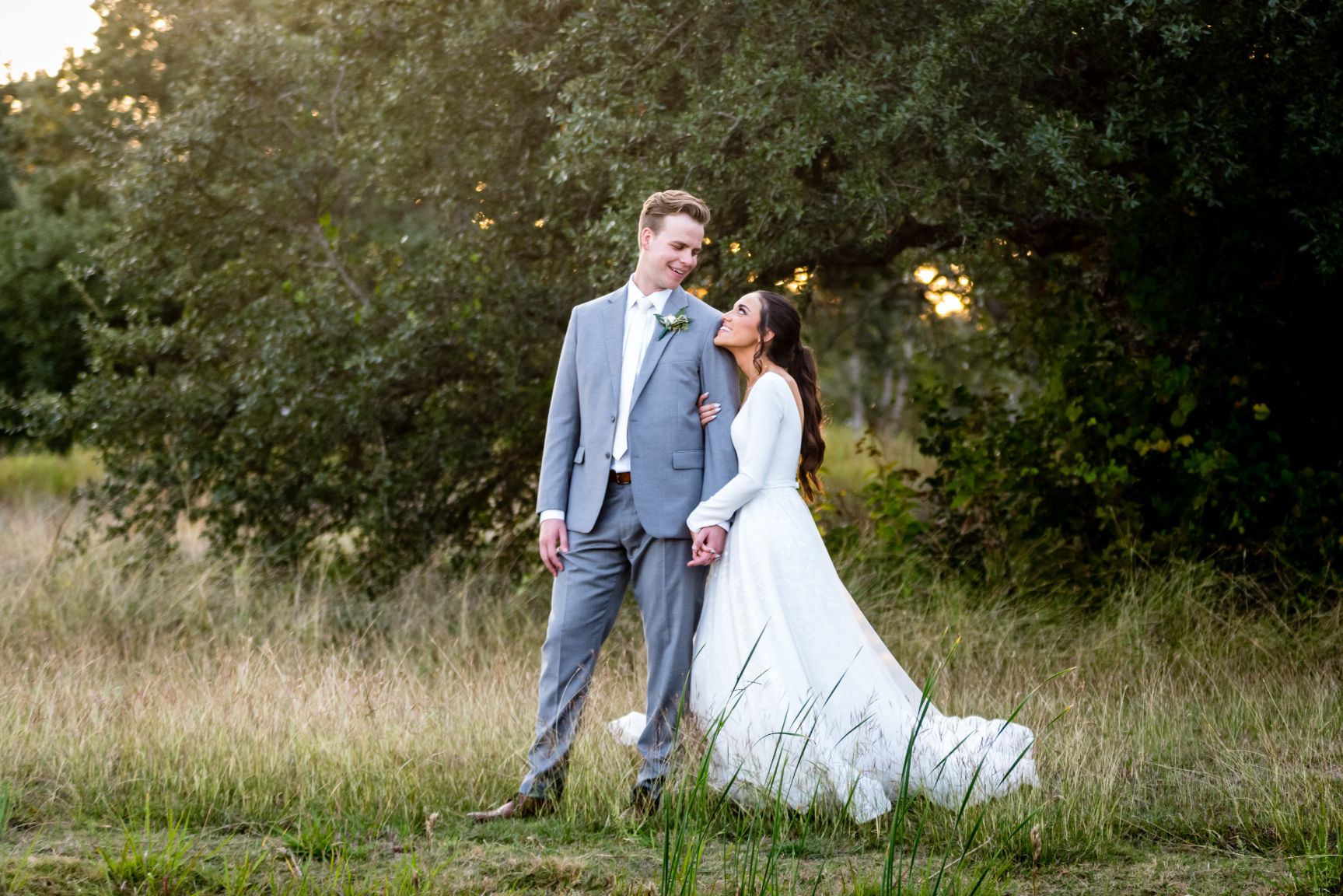 Bride wearing white dress with long sleeves holds hands with groom wearing gray suit in front of large green oak trees. 
