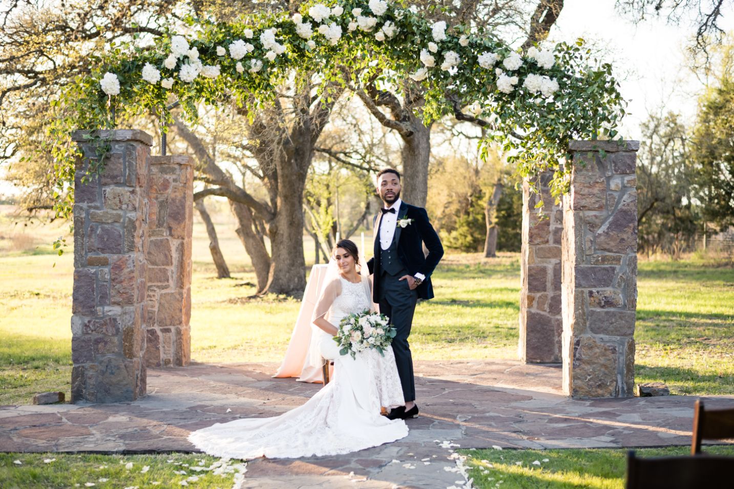 A bride sits in a chair with groom touching her shoulder and looking off into the distance. The stone towers are in the background, with a warm sunset lighting the oak strees.