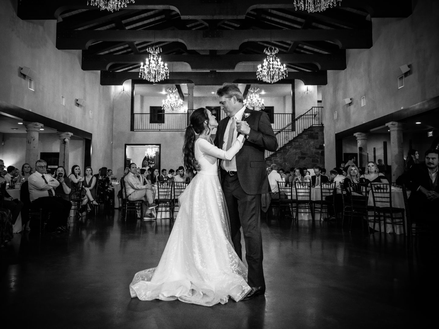 A bride dances with her father in a large banquet style hall. Wood beams on the ceiling with chandeliers. 