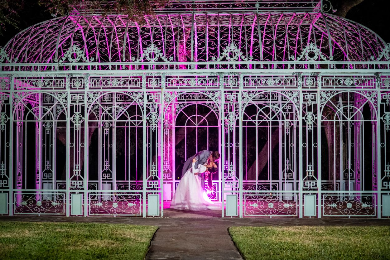 Bride and groom kiss under a green metal gazebo at night with pink lights illuminating the structure. 