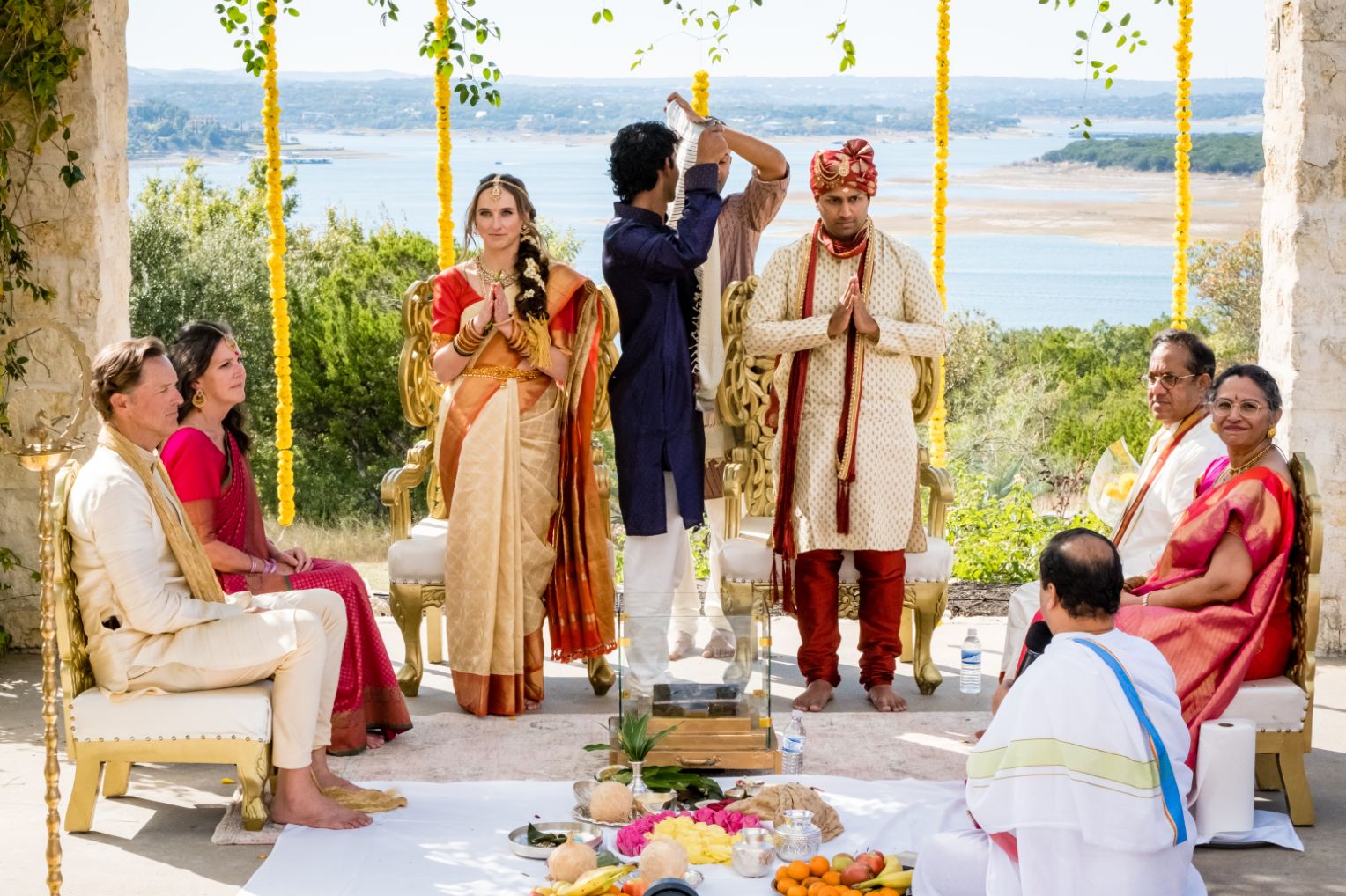 Bride and groom wearing traditional Indian garments stand with prayer hands during Indian wedding ceremony at Vintage Villas. The family members are seating around the couple with Lake Travis views in the background.