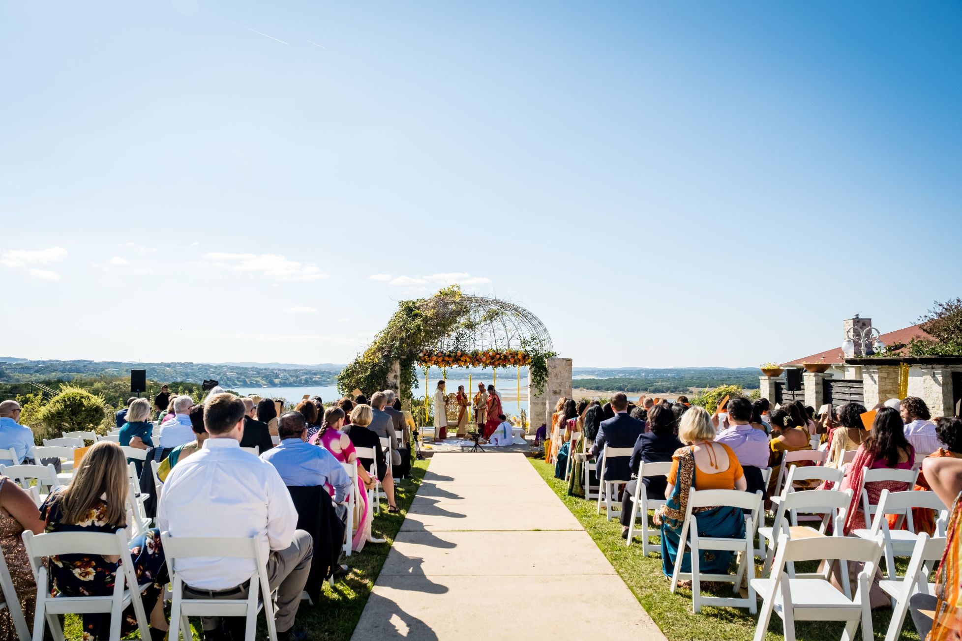 Indian wedding ceremony at Vintage Villas with bright blue sky in the background and Lake Travis views in the distance. Wedding ceremony guests are seated in rows on white lawn chairs.