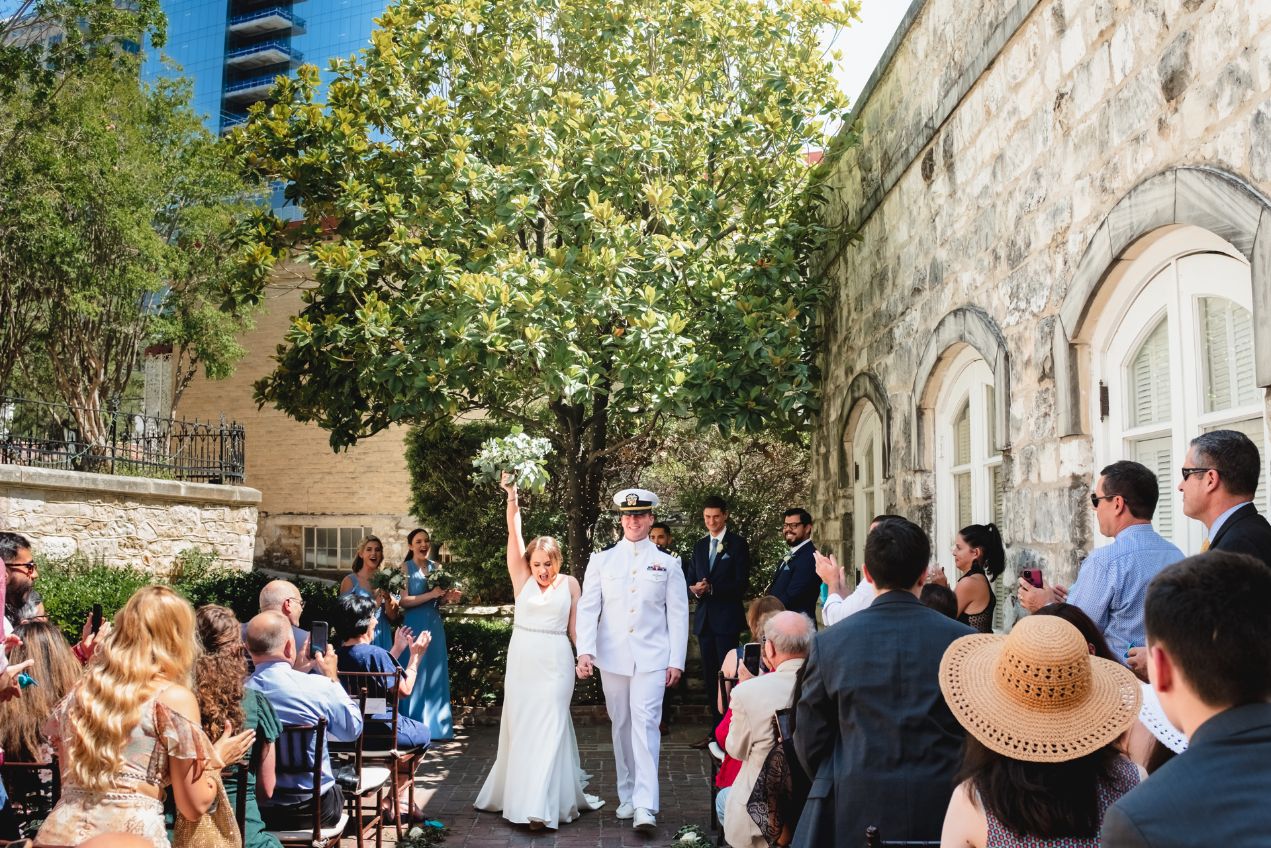 Brides holds grooms hands with her other hand up in the air in celebration as the couple walk down the aisle of their wedding ceremony at Chateau Bellevue. Tall downtown buildings are seen in the back behind a set of trees in a romantic outdoor courtyard with stone building facade.