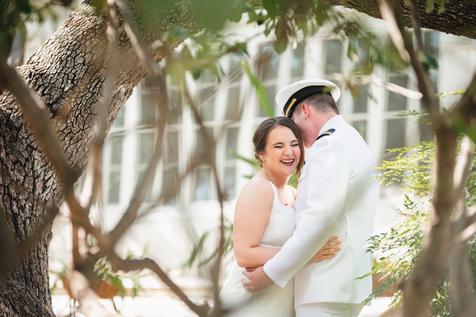 Bride and groom hold each other under a tress. The groom is wearing a white military uniform, the bride is smiling. 
