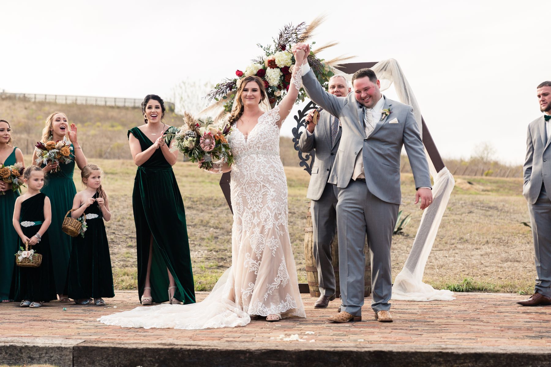 Bride and groom stand hold hands above their heads near a hexagon shaped wedding altar with wood barrel during wedding ceremony at Two Wishes Ranch in Austin Texas.