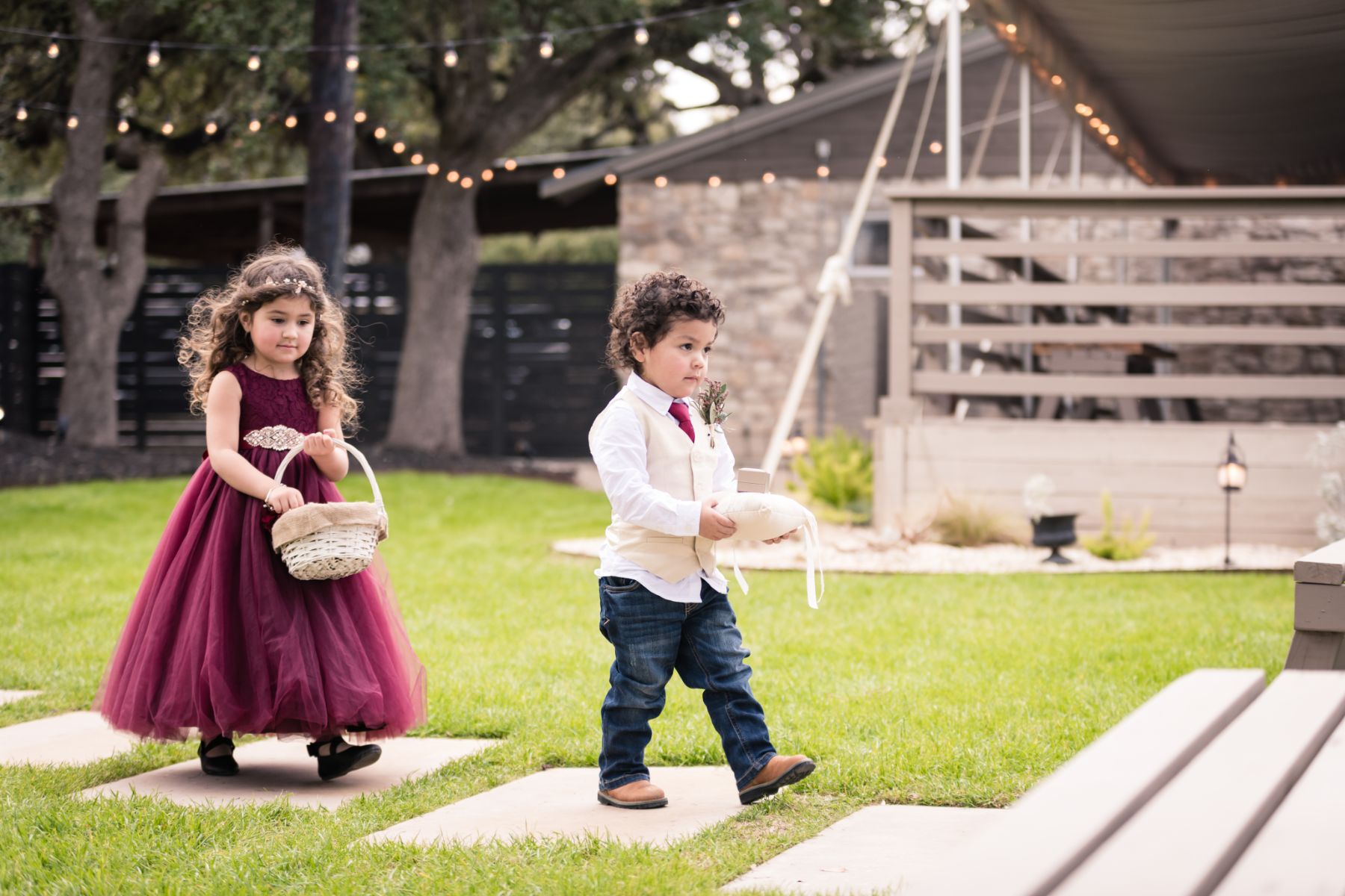 Flower girl, wearing maroon dress and ring bearer, walk down stone steps on a green grass lawn of an outdoor ceremony space at Stonehouse Villa.
