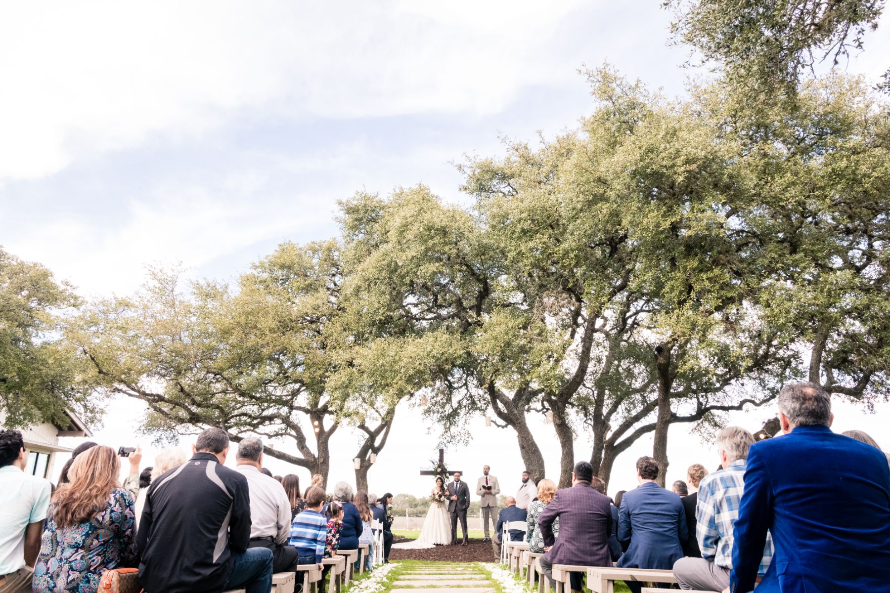 A vride and groom stand beneath Texas oak trees with their ceremony guests seated on benches at Stonehouse Villa.